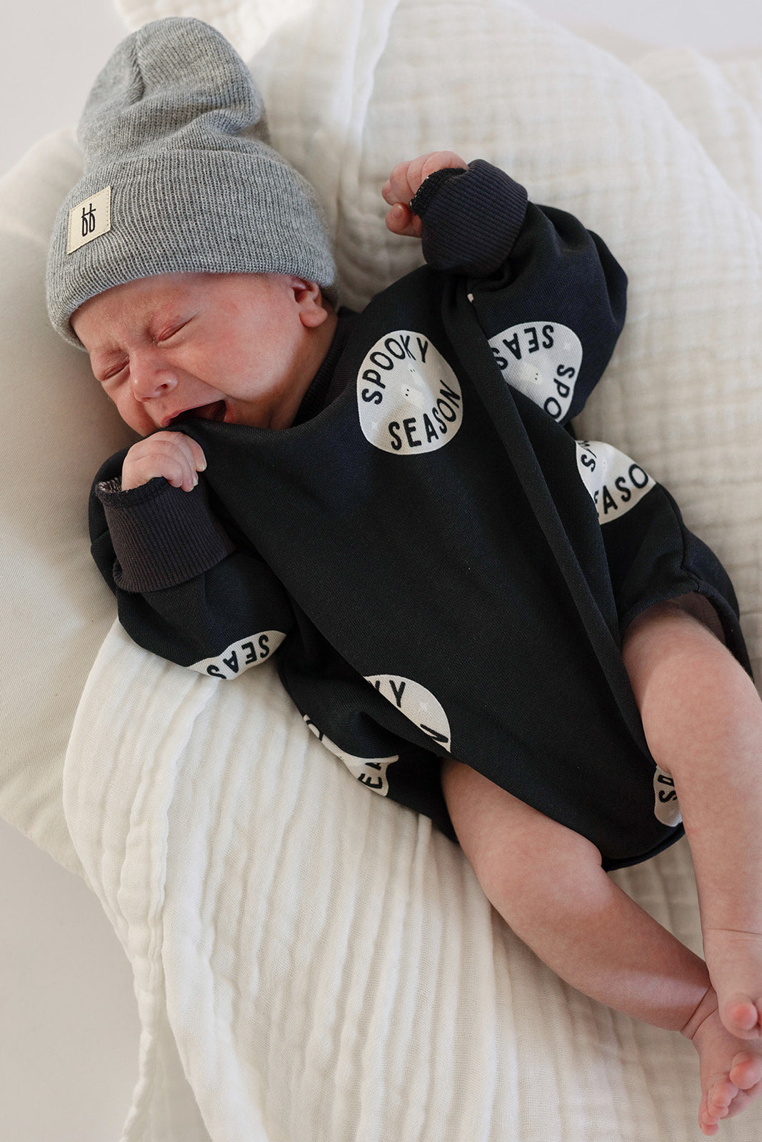 A newborn wearing the forever french baby Sweatshirt Romper | Spooky Season lies on a soft white blanket, crying and clinging to the cozy fabric—the perfect Halloween outfit for your little one.