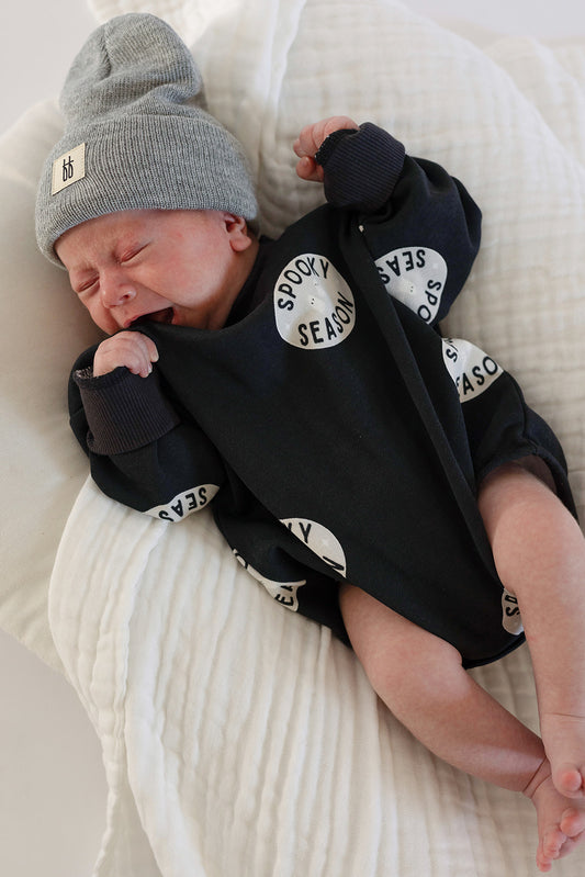 A newborn wearing the forever french baby Sweatshirt Romper | Spooky Season lies on a soft white blanket, crying and clinging to the cozy fabric—the perfect Halloween outfit for your little one.