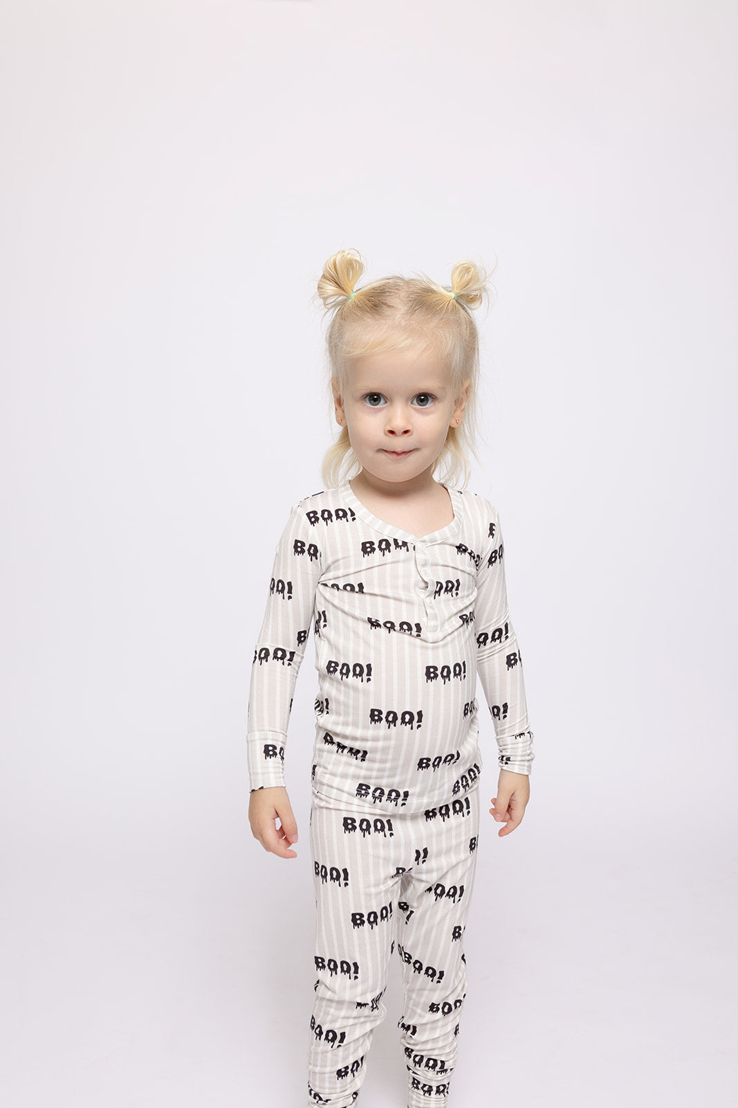 A young child with blonde hair in two buns stands against a white background, wearing forever french baby Bamboo Two Piece Pajamas | Boo!—hypoallergenic kids sleepwear featuring a playful "ROAR!" dinosaur print and smiling at the camera.