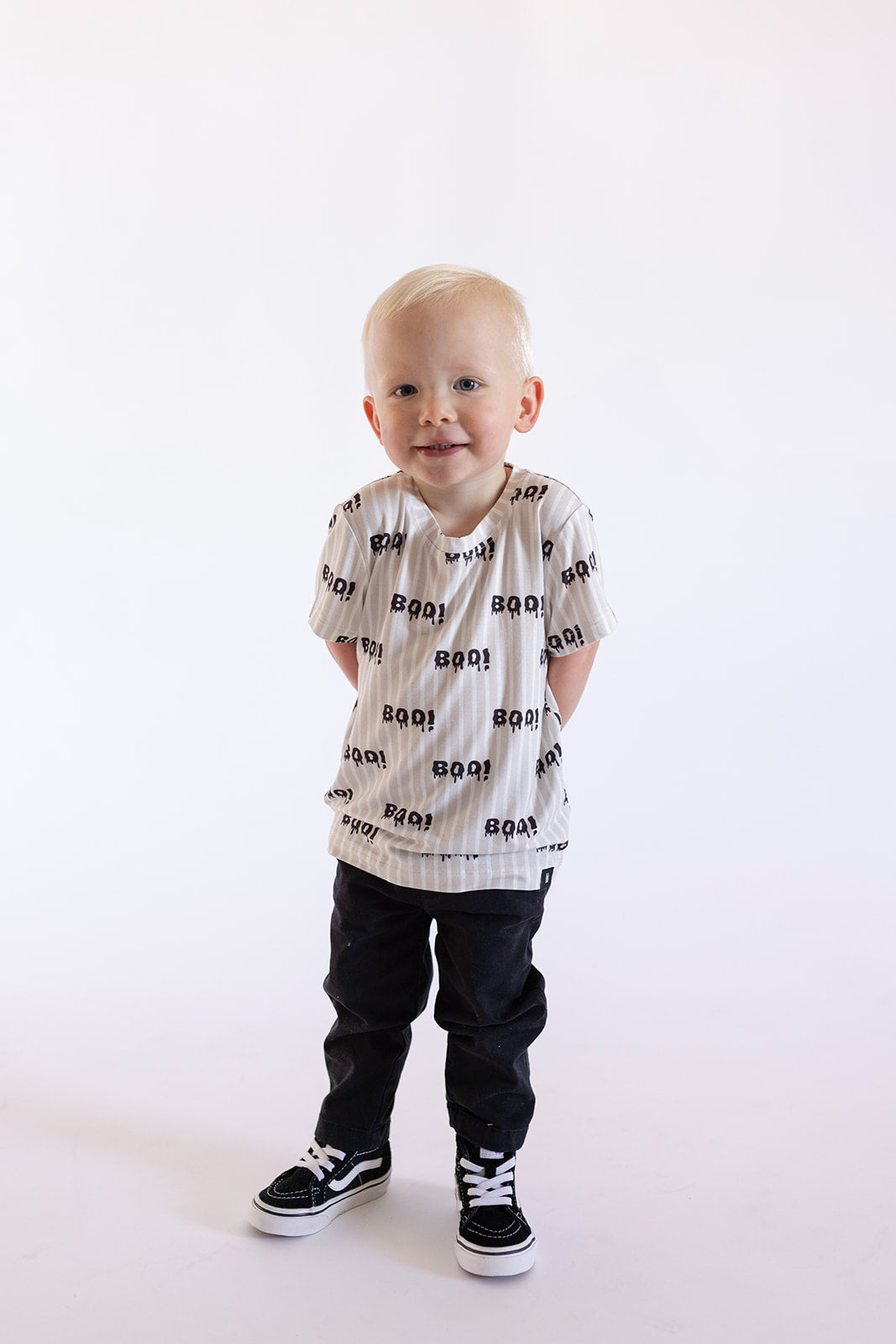 A young child with short blond hair smiles in a forever french baby "Child Bamboo T-Shirt | Boo!"—a white bamboo kids tee with black text—paired with dark pants and black-and-white sneakers, against a plain light background.