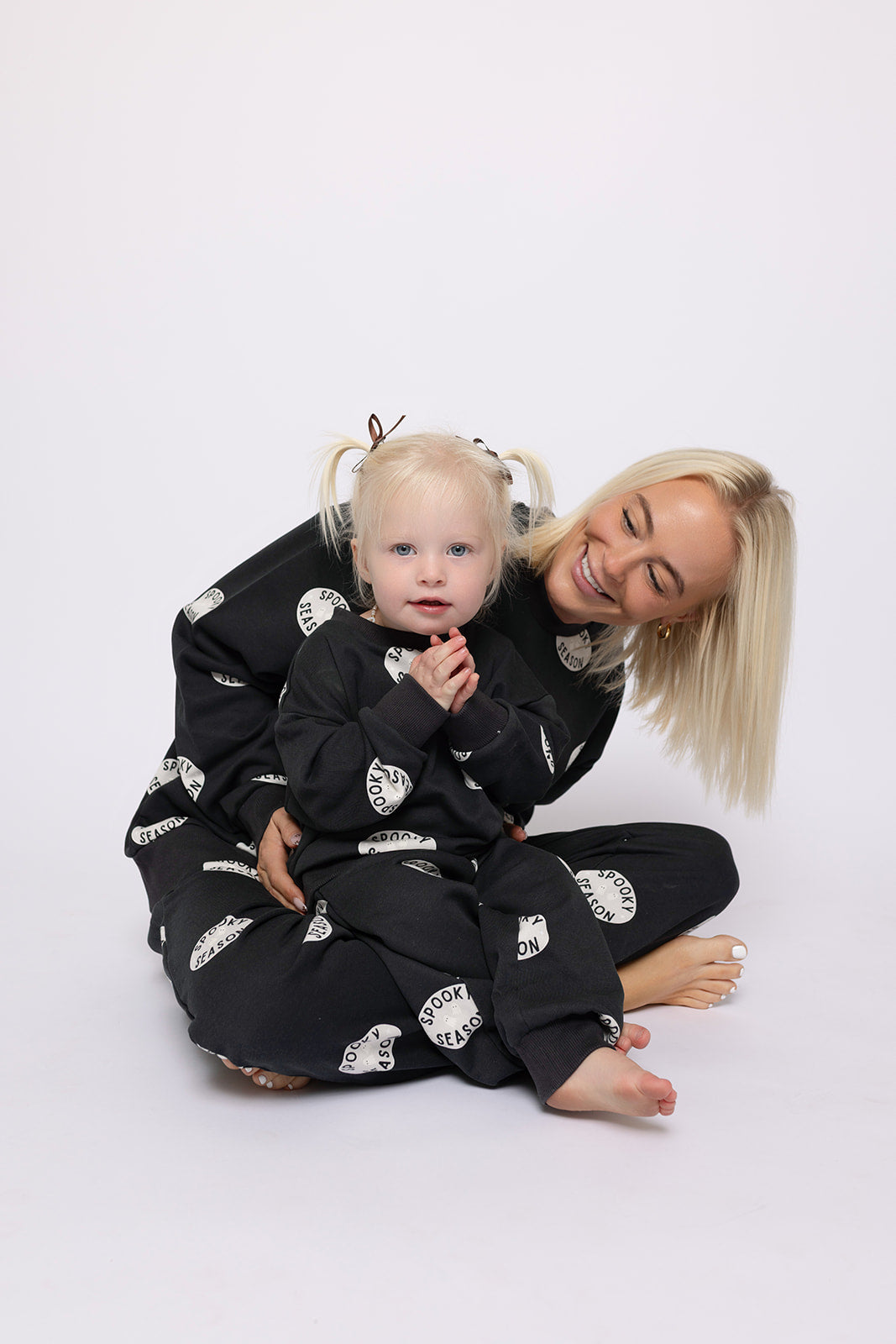 A smiling woman hugs a young child with blonde pigtails, both wearing forever french baby’s Child Sweat Set | Spooky Season in black with white circles—an ideal matching look for Halloween, set against a plain white background.