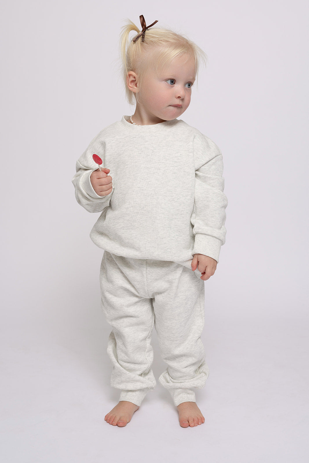 A young toddler in the forever french baby Child Sweat Set | Heather Grey stands barefoot on a white background, holding a red lollipop.
