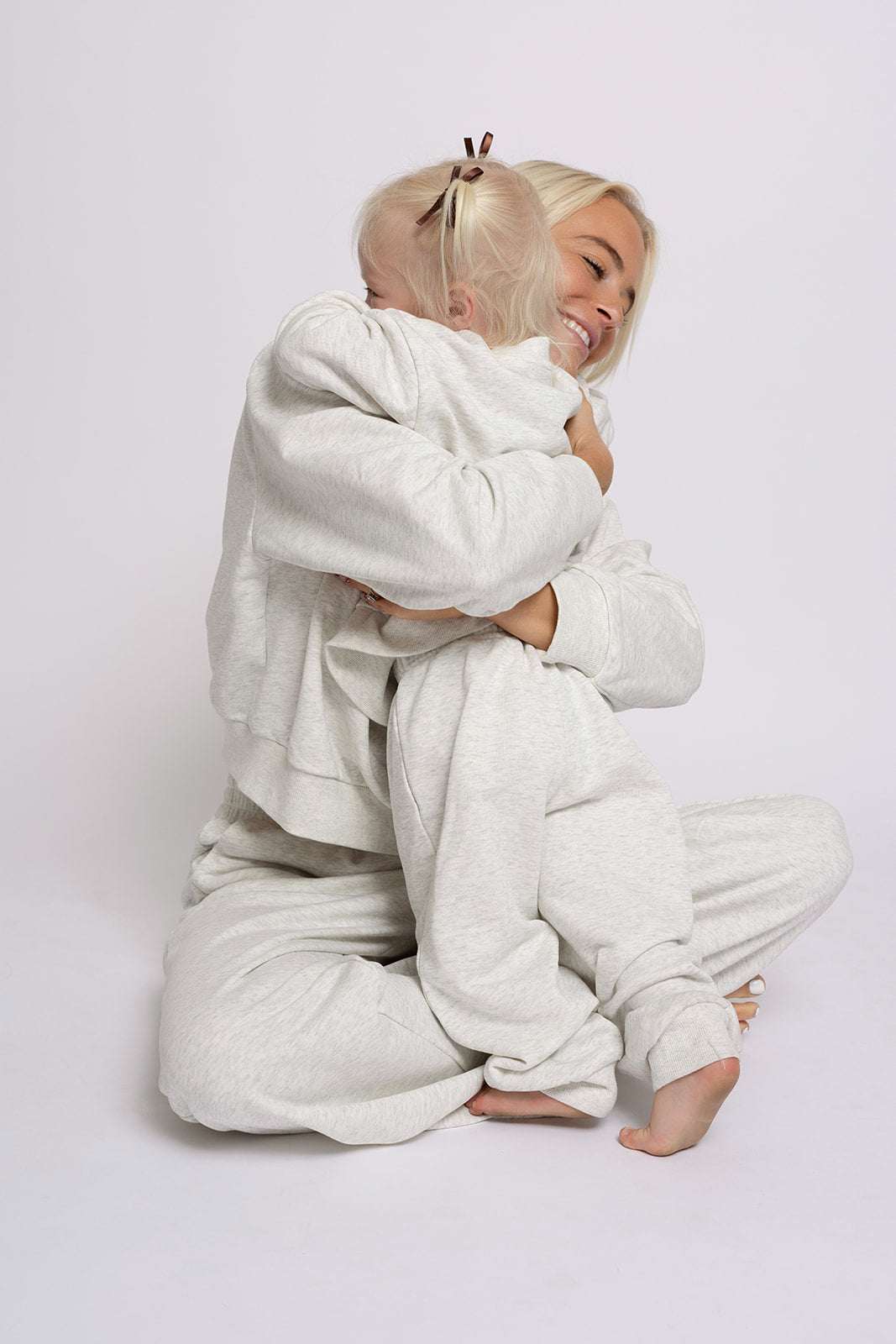 A woman in a light gray sweat set hugs a smiling child dressed in the forever french baby "Child Sweat Set | Heather Grey," both in matching family outfits against a plain white background.