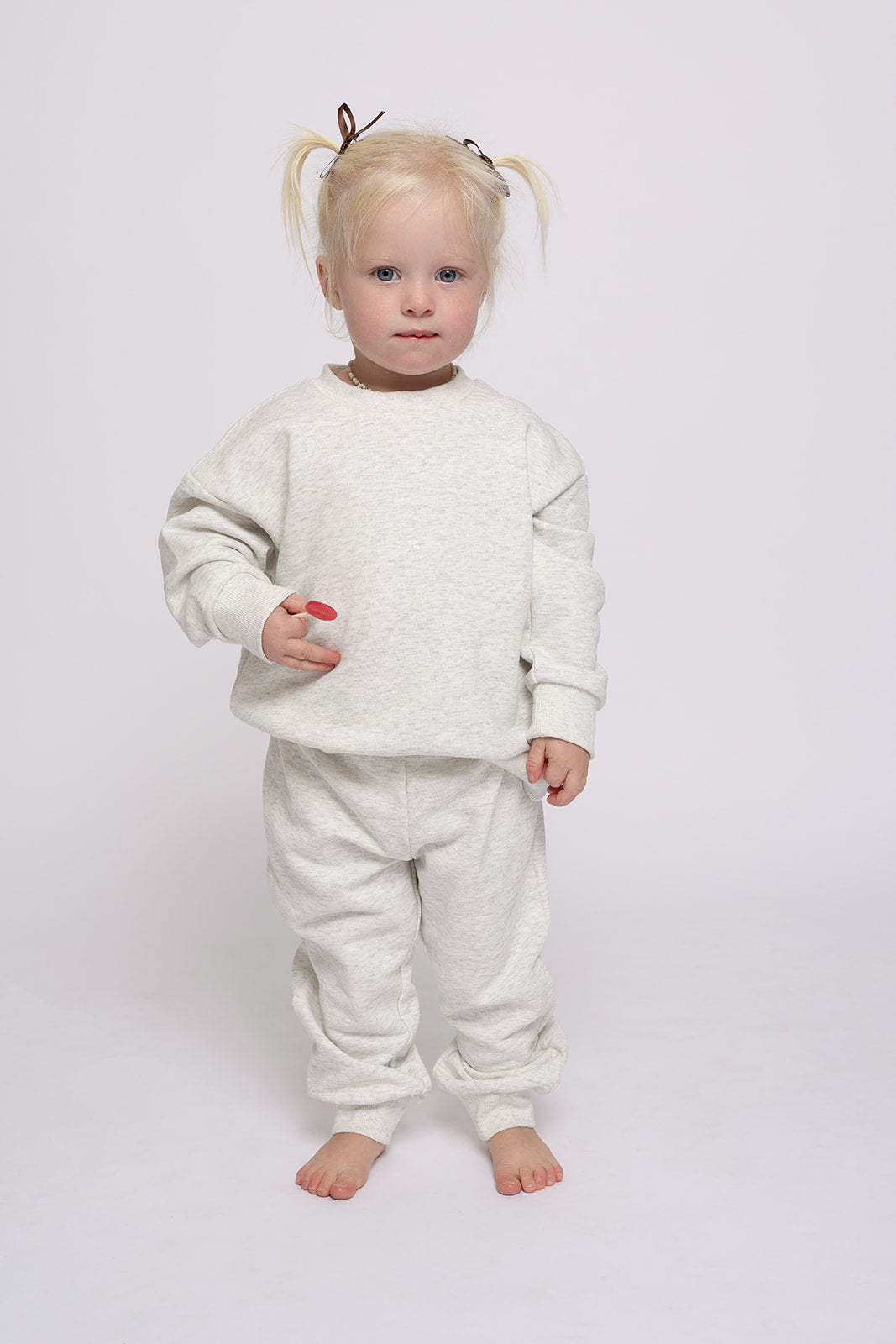 A young blonde toddler with pigtails stands barefoot on a white background, wearing the forever french baby Child Sweat Set in Heather Grey, looking directly at the camera.