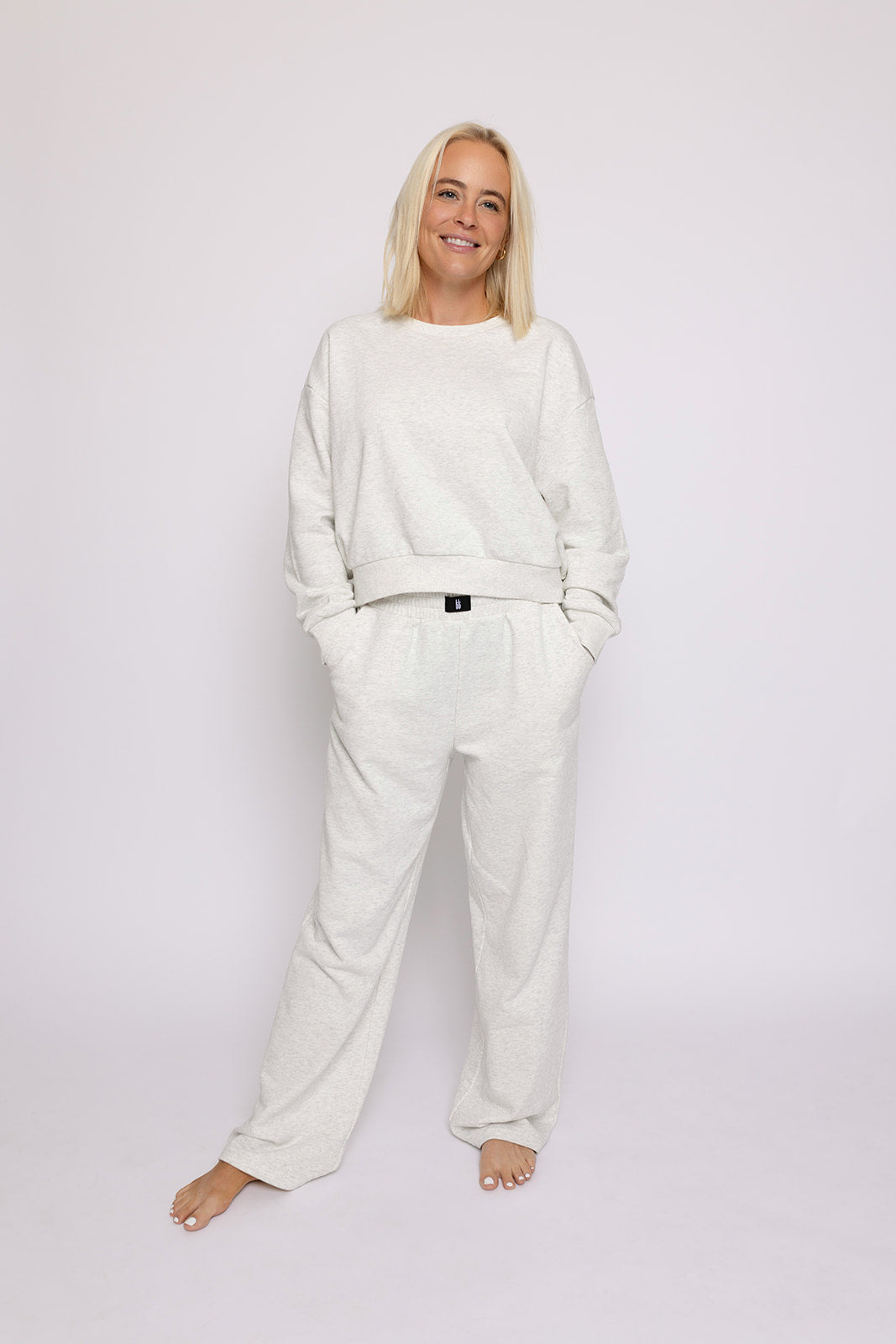 A blonde woman stands barefoot against a white background, smiling, wearing the forever french baby Women's Sweat Set in Heather Grey—featuring a loose-fit sweatshirt and matching sweatpants with her hands in her pockets.