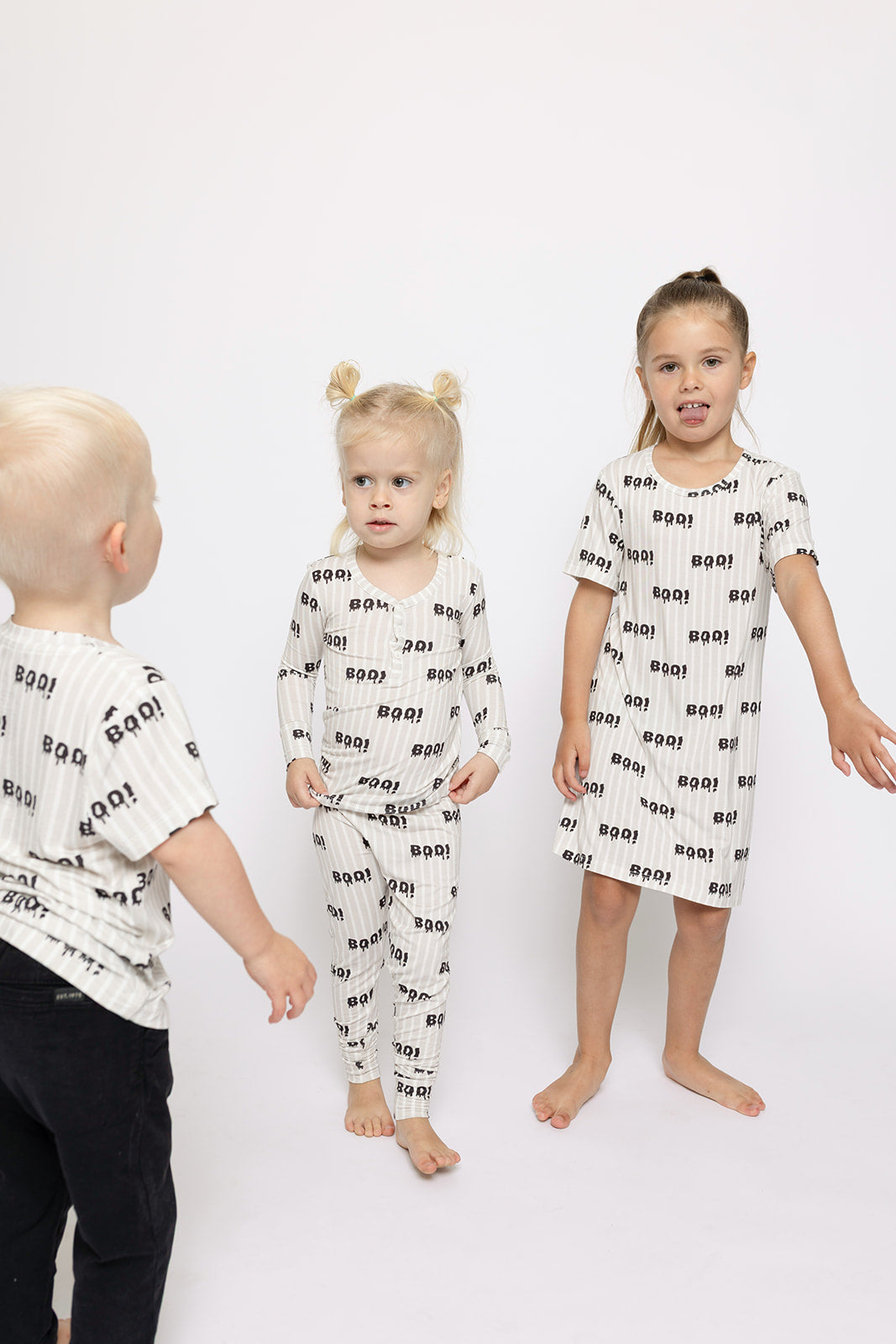 Three young children wear matching forever french baby Bamboo Two Piece Pajamas | Boo!, featuring “Boo!” in black on white fabric. The child on the right sports a dress, while the others wear long-sleeve tops and bottoms.