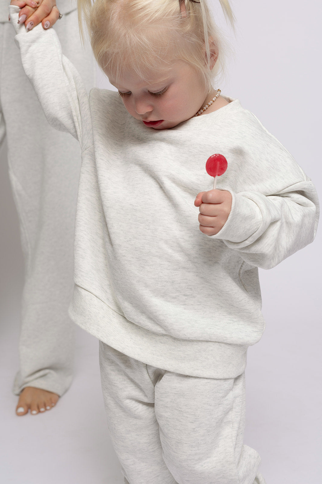 A young blonde child wears the forever french baby Child Sweat Set in Heather Grey, holding a red lollipop and looking down while standing barefoot next to an adult against a plain white background.