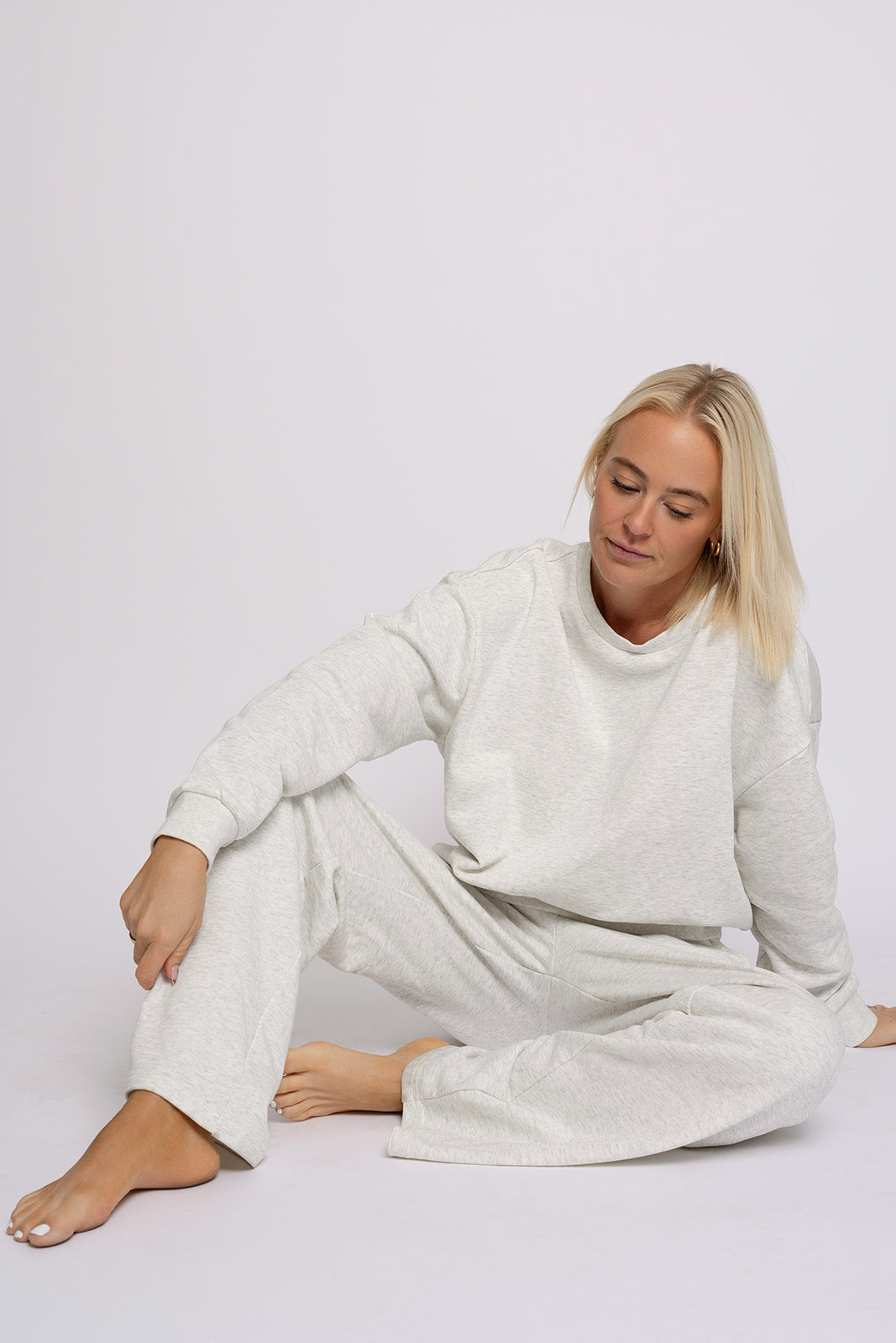 A woman with blonde hair sits against a plain background in the forever french baby Women's Sweat Set | Heather Grey, looking relaxed with one leg bent and the other extended.