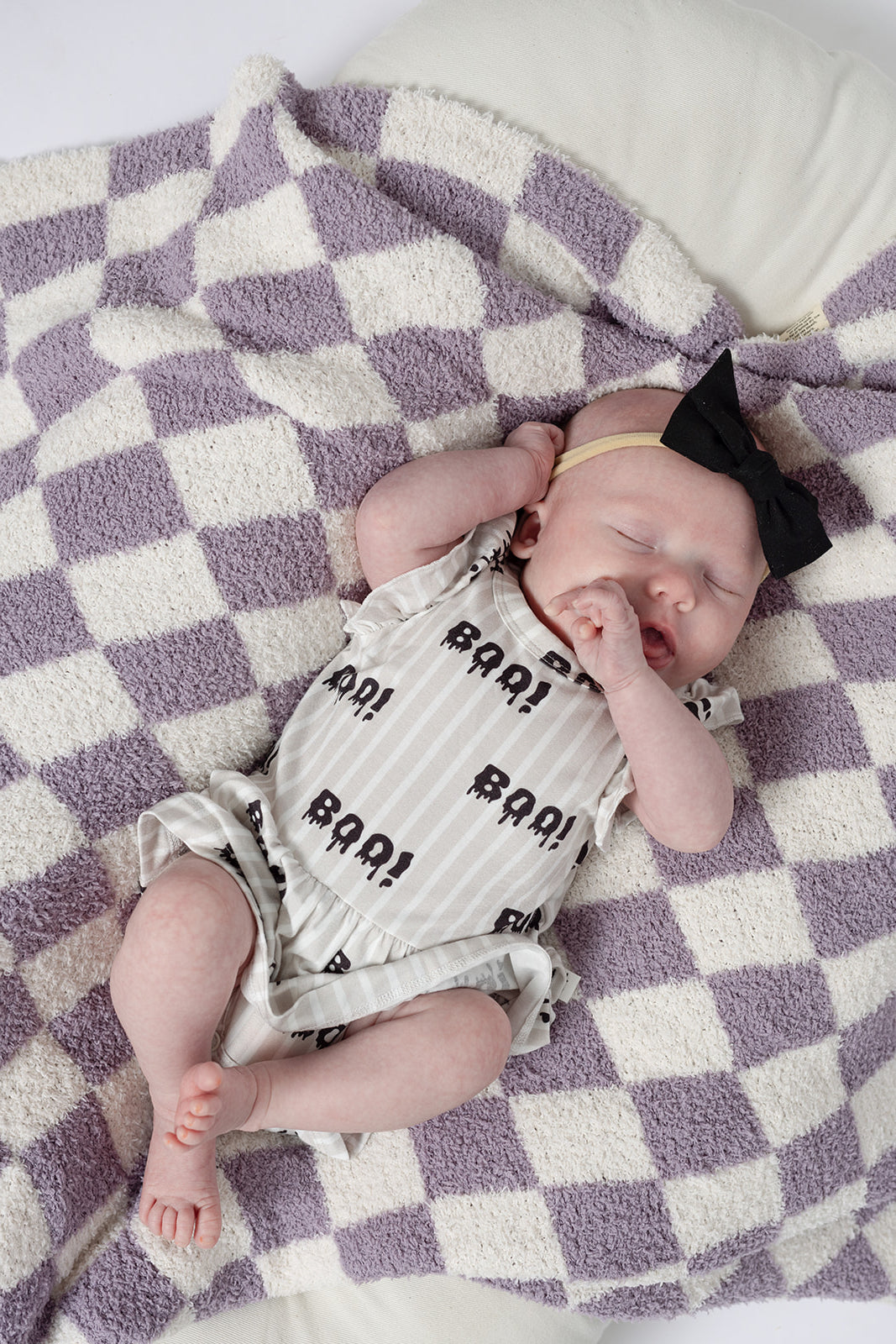 A baby in forever french baby's Bamboo Ruffle Romper | Boo! with a large black bow headband lies yawning on a soft, white and lavender checkered blanket. The hypoallergenic bamboo romper ensures cozy comfort.