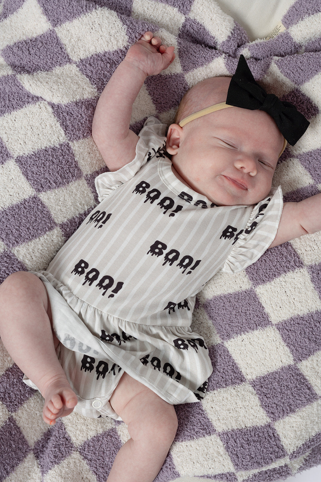A baby in a forever french baby Bamboo Ruffle Romper | Boo! with a black bow headband sleeps peacefully on a purple and white checkered blanket, one arm raised above their head.