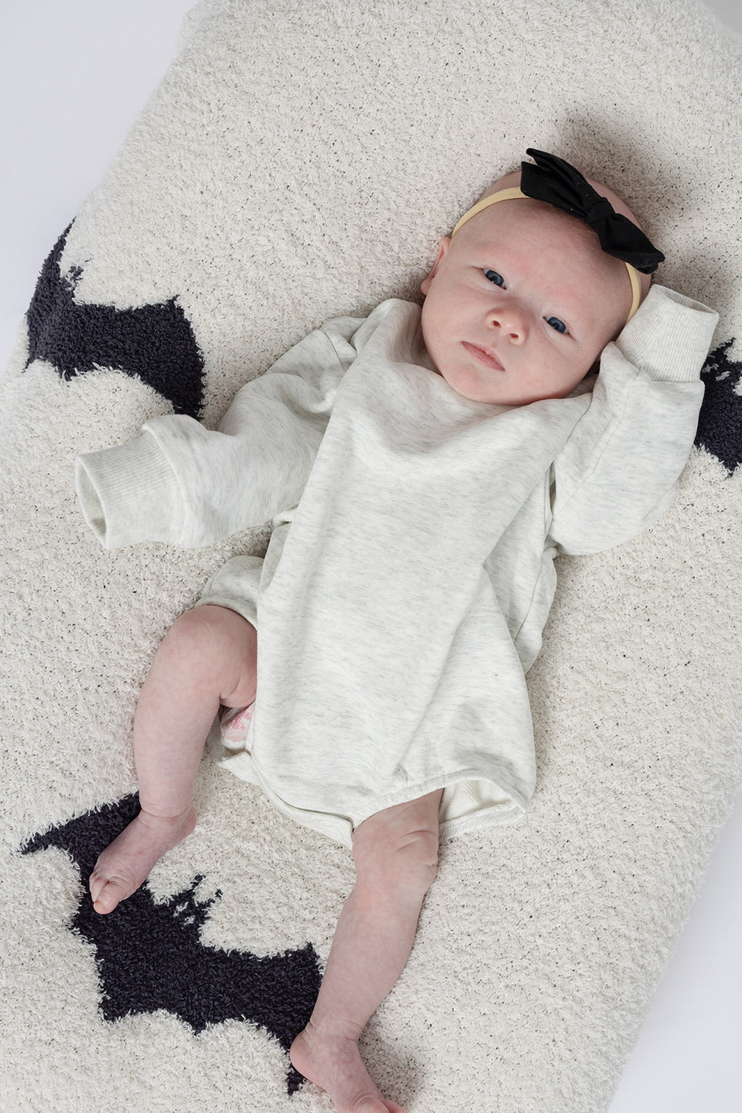 A newborn wearing the forever french baby Sweatshirt Romper in Heather Grey and a black bow headband lies on a cream blanket with black bat shapes, gazing upward with one arm raised beside their head.