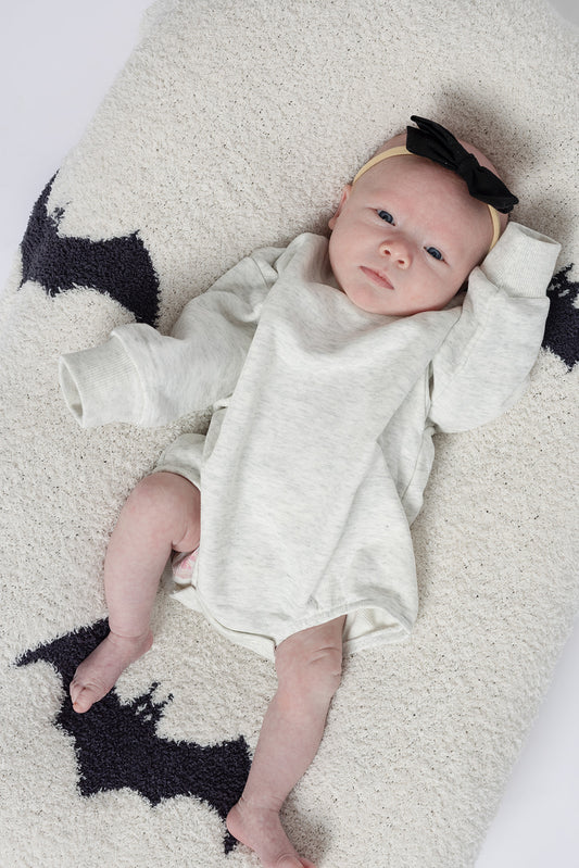 A newborn wearing the forever french baby Sweatshirt Romper in Heather Grey and a black bow headband lies on a cream blanket with black bat shapes, gazing upward with one arm raised beside their head.