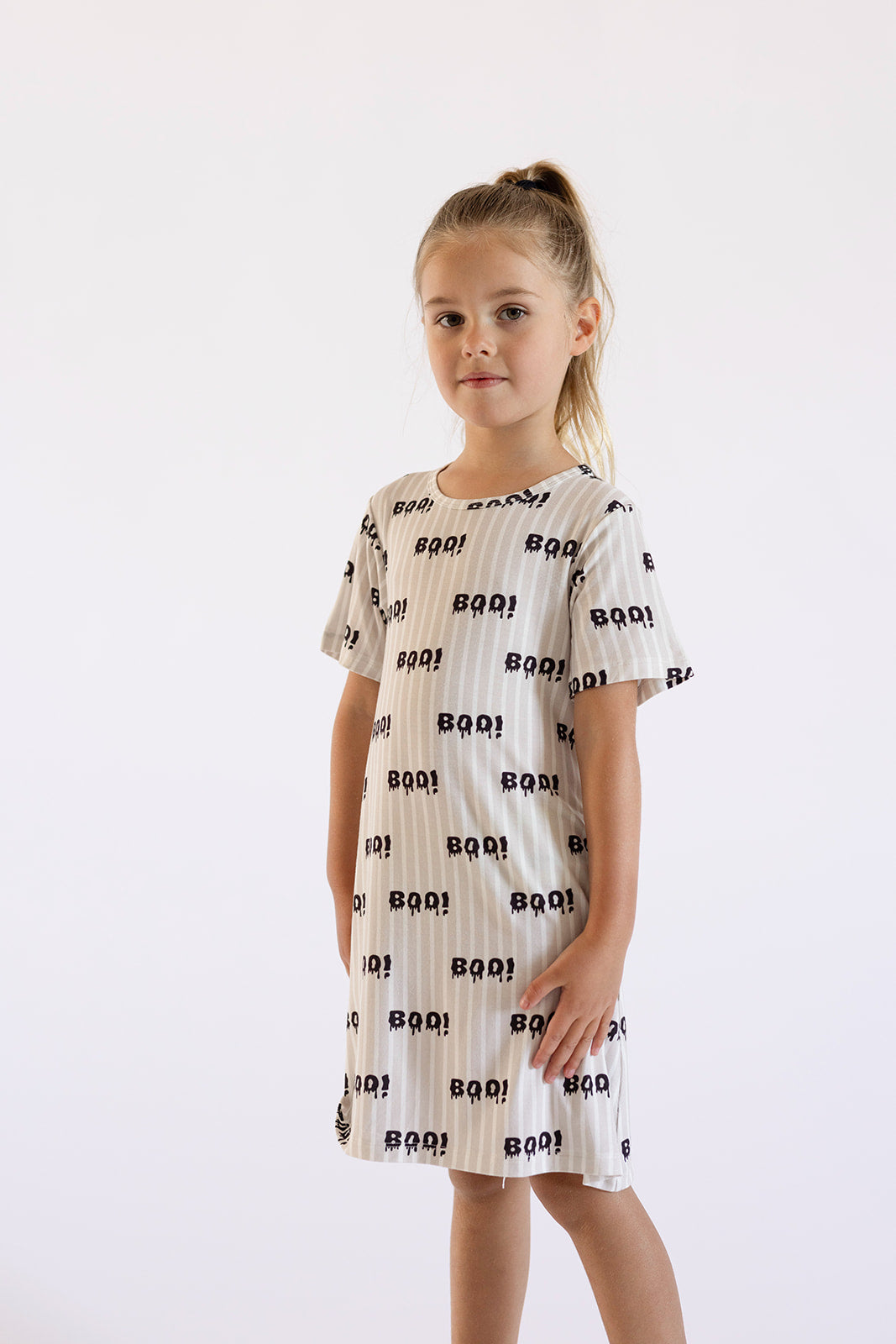 A young girl with light brown hair in a ponytail stands against a white background, wearing the forever french baby Bamboo T-Shirt Dress | Boo! featuring black "BOO!" text and small ghost illustrations.