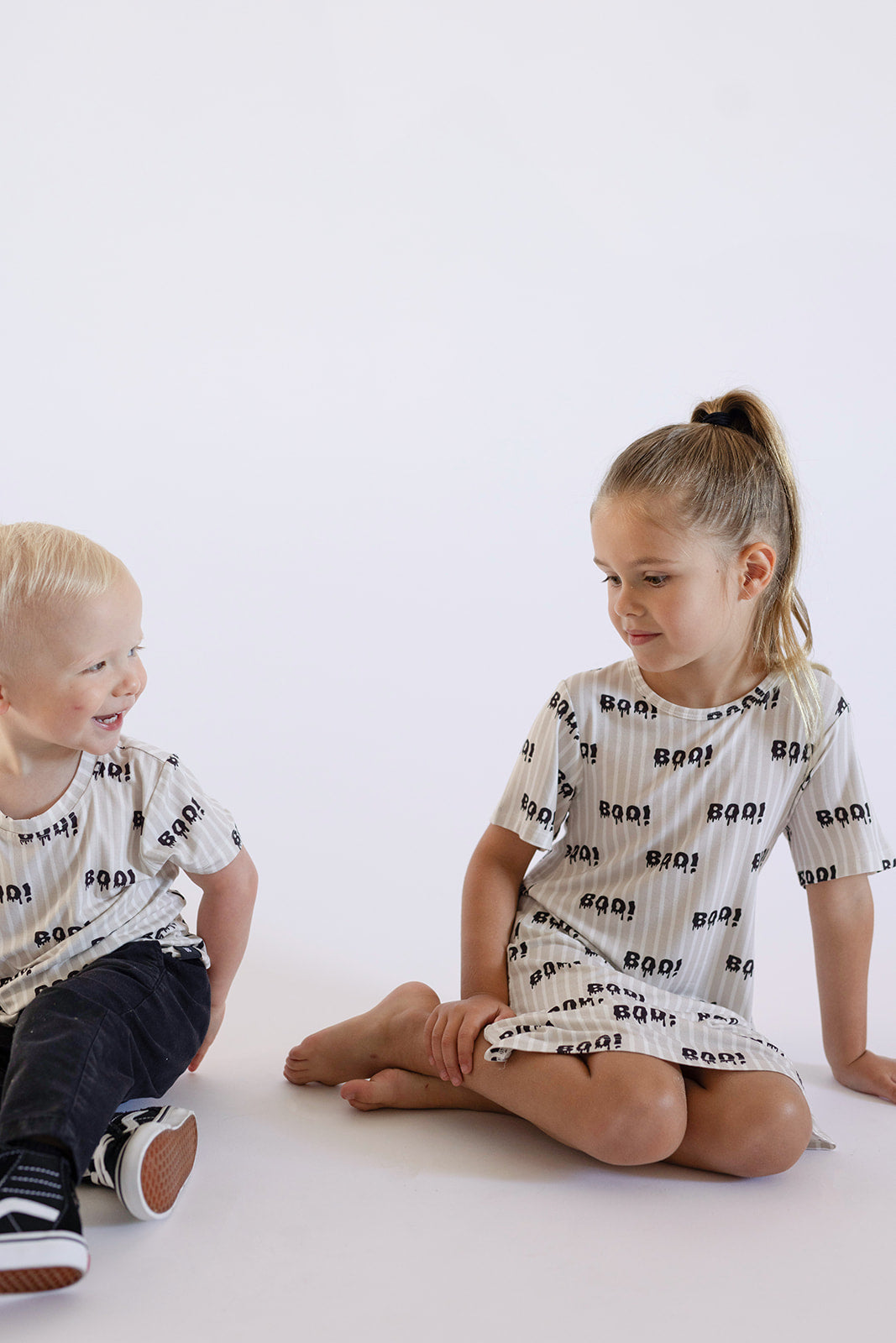 Two young children sit against a white background. The boy wears a comfy tee, while the girl—hair in a ponytail—sports the hypoallergenic Bamboo T-Shirt Dress | Boo! by forever french baby, showcasing soft bamboo clothing for kids.