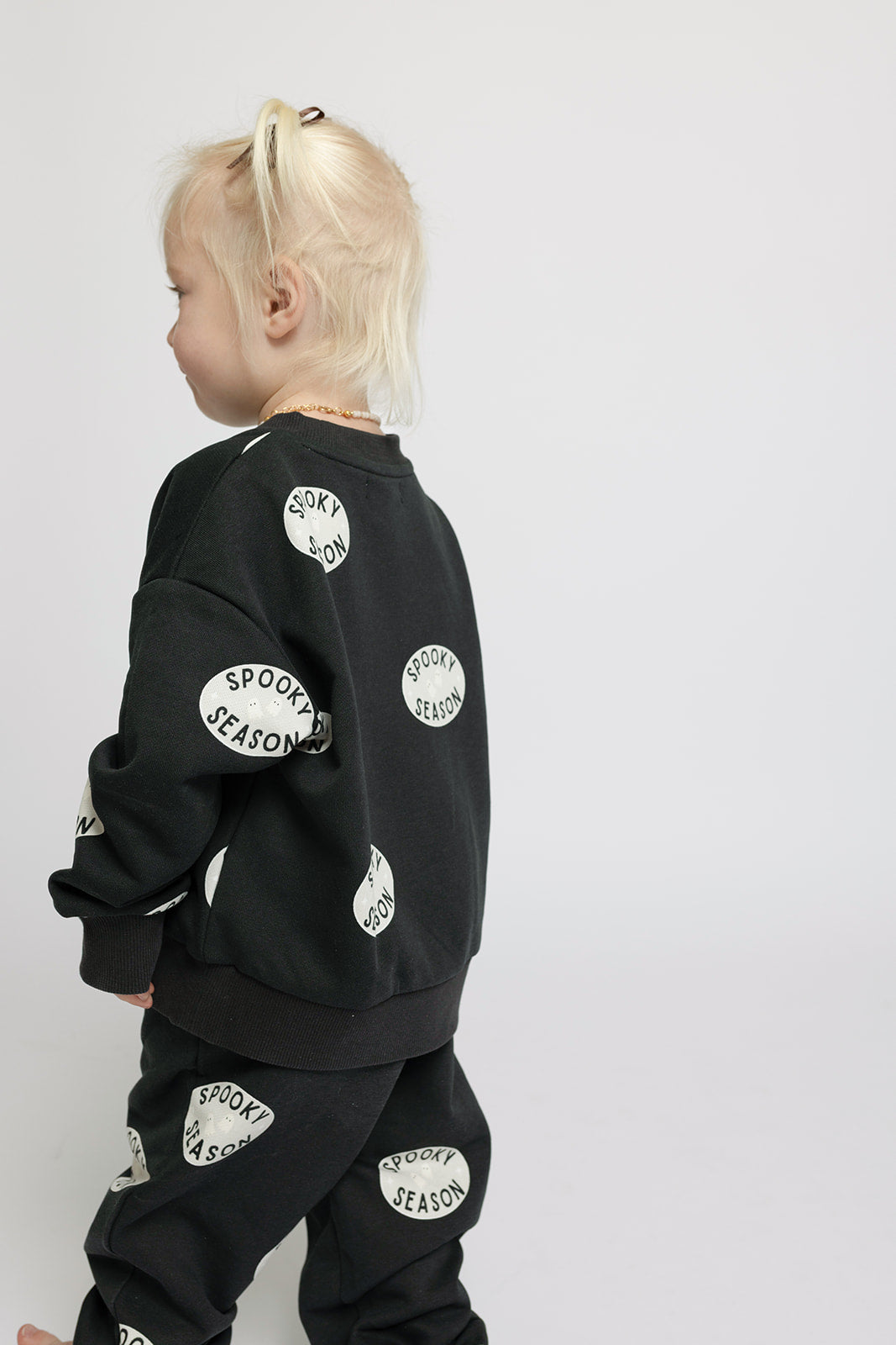 A toddler with blonde pigtails stands sideways on a plain white background, wearing the forever french baby Child Sweat Set | Spooky Season featuring black pants and top with white “SPOOKY SEASON” patches.
