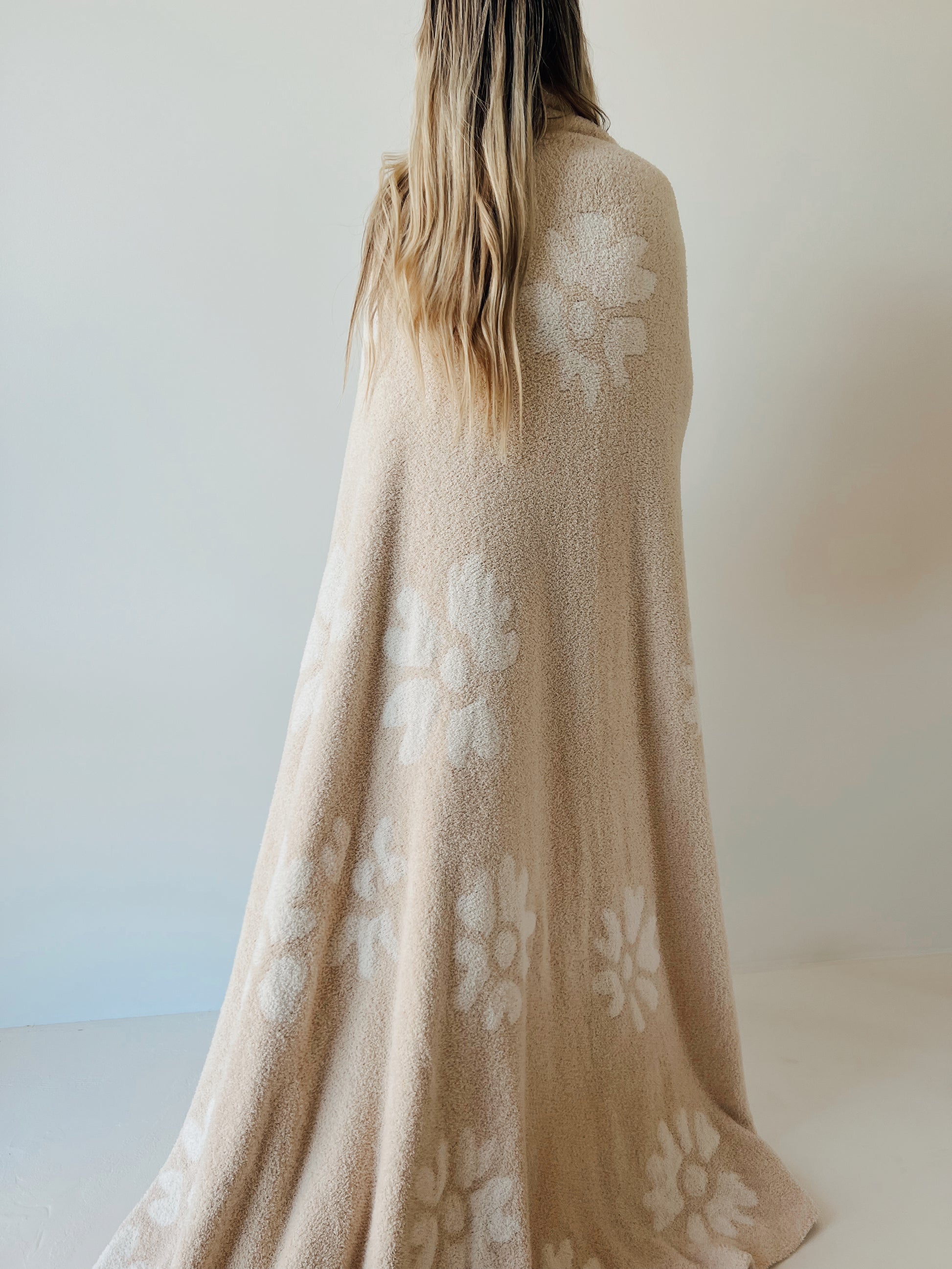 A person with long, wavy hair, seen from behind, draped in the Plush Blanket | Lazy Daisy by forever french baby. The blanket, made of velvety-rich textured microfiber feather yarn and certified by OEKO Standard 100, is adorned with white floral patterns and stands out against a plain light-colored background.