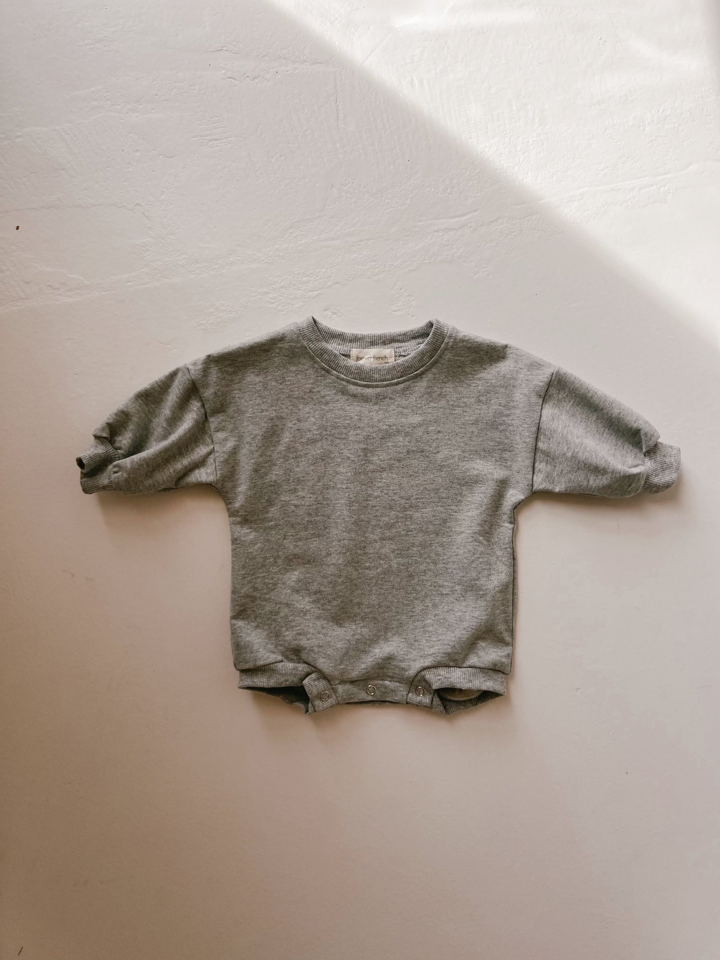 A small, gray Sweartshirt Romper | Stormi from forever french baby is laid out flat against a light-colored background. The romper features a crew neckline, ribbed cuffs on the sleeves, and snap closures at the bottom for easy diaper changes. It's crafted from the softest material for ultimate comfort.