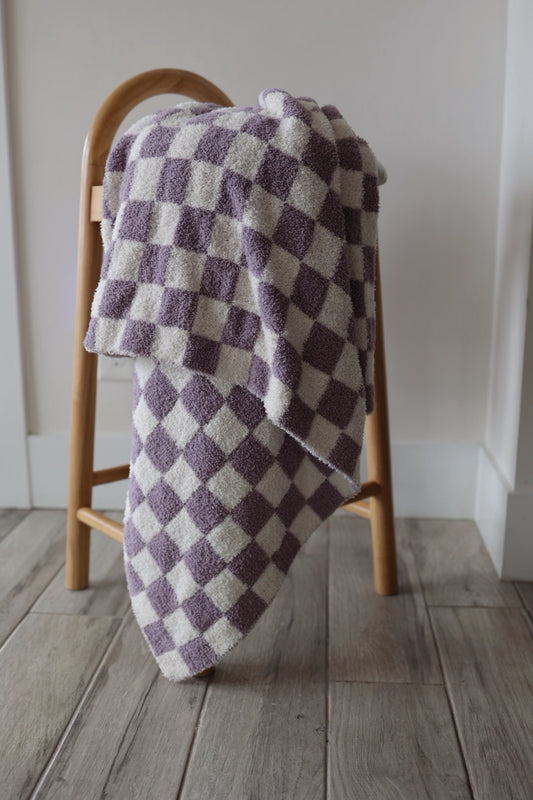 A wooden stool with the forever french baby Plush Blanket | Grape Soda, a soft purple and white checkered throw, draped over it sits on a light wooden floor against a plain white wall.