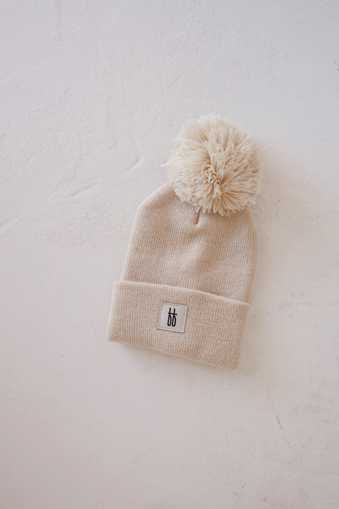 The ff Pom Top Knit Beanie in oatmeal from forever french baby is lying on a white textured surface. Featuring a large, stylish pom on top and a folded brim adorned with a small rectangular fabric tag showcasing a simple black and white design, this winter hat is both cozy and chic.