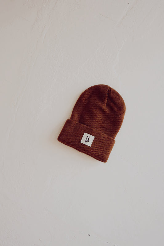 The ff Knit Beanie in Chestnut by forever french baby, featuring a folded brim and a small white rectangular patch with a minimalist logo, is placed against a textured off-white background.