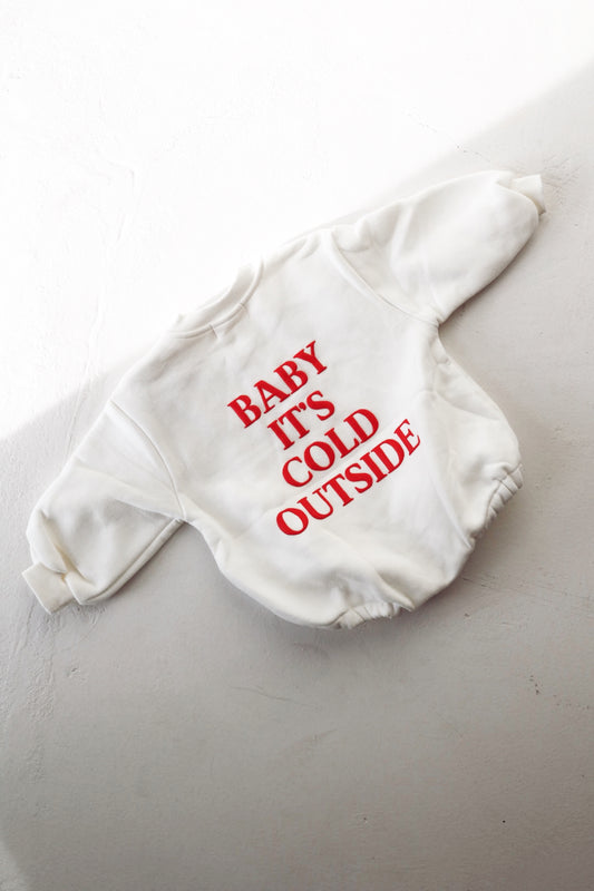 A white Sweatshirt Romper from forever french baby, featuring red text that reads "BABY IT'S COLD OUTSIDE," is displayed on a white surface, illuminated by side sunlight. Ideal for chilly weather attire, it effortlessly complements family matching outfits, adding cozy charm for everyone.