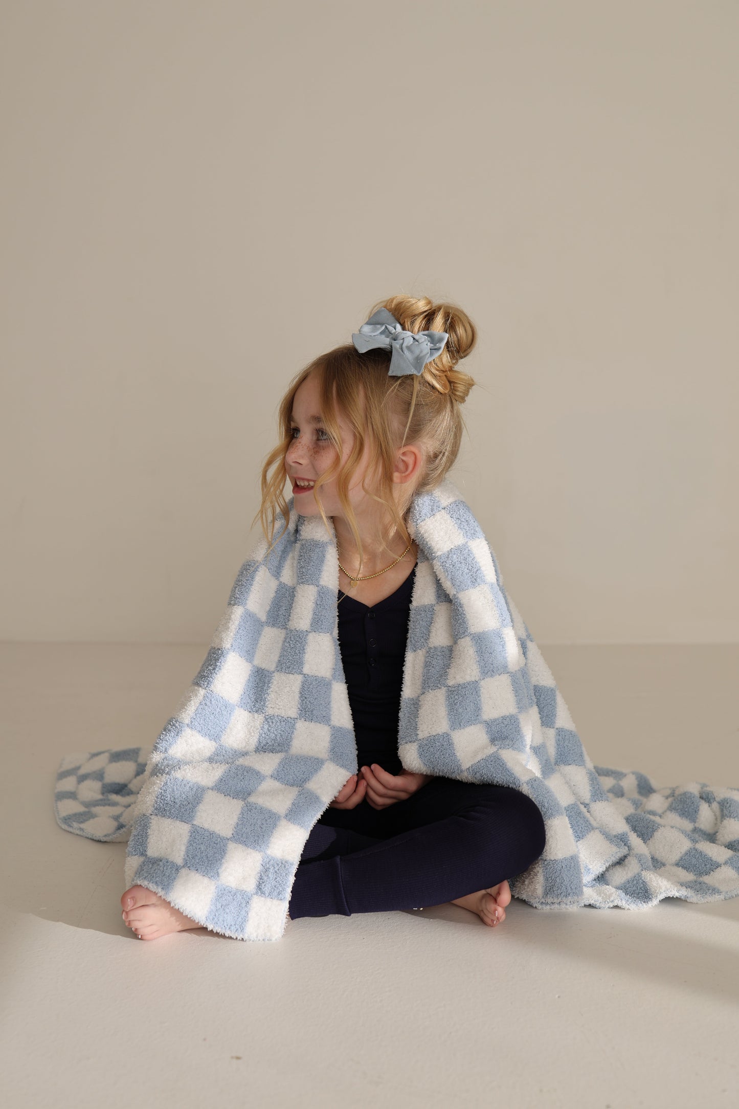 A young blonde girl with a bun, blue bow, and navy outfit sits on the floor wrapped in the forever french baby Plush Blanket | Morning Check, an Oeko-Tex certified light blue and white checkered blanket, smiling to the side.