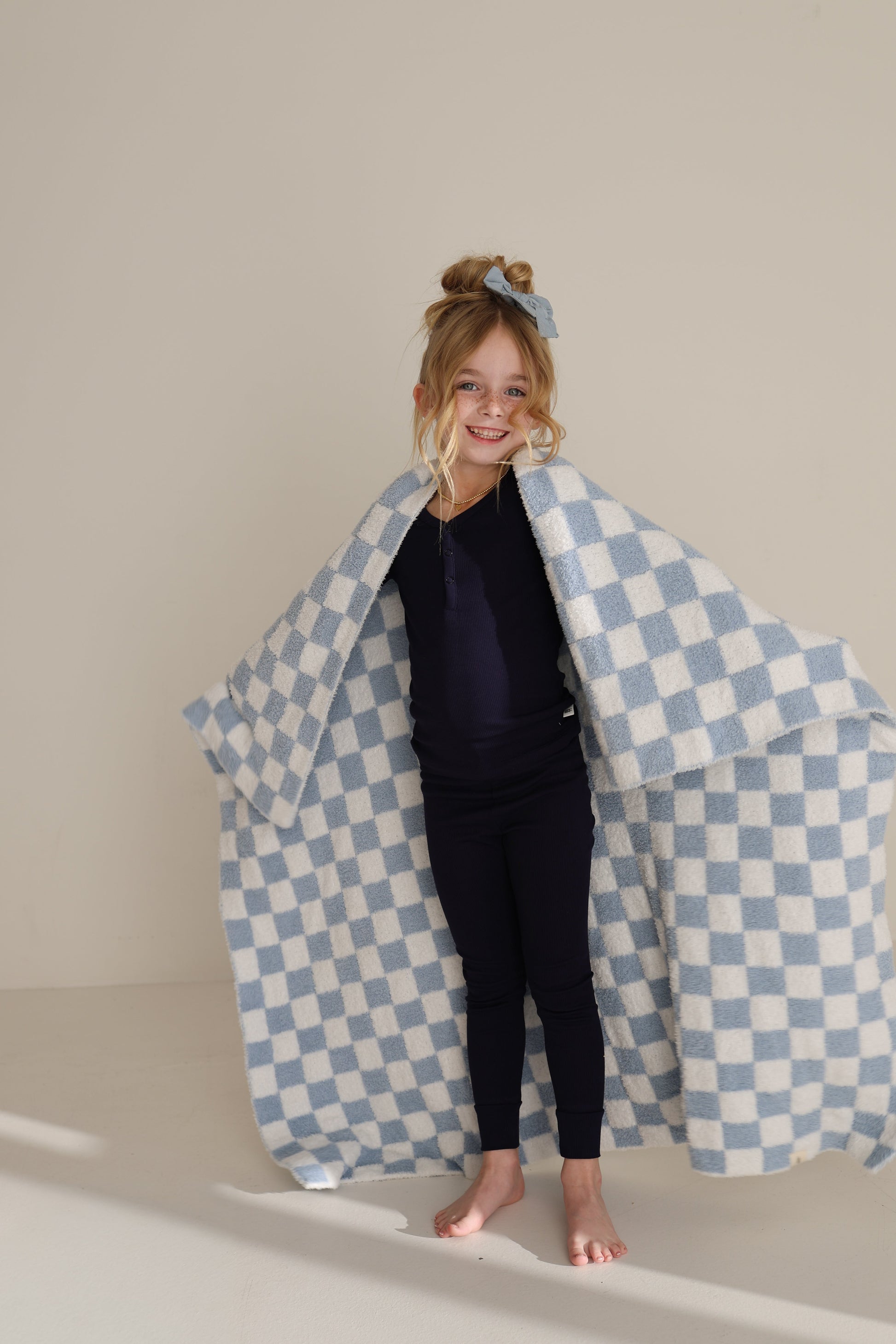 A smiling young girl with a blue bow stands barefoot, wrapped in the forever french baby Plush Blanket | Morning Check, featuring blue and white checks and Oeko-Tex certified microfiber, against a plain light-colored wall.