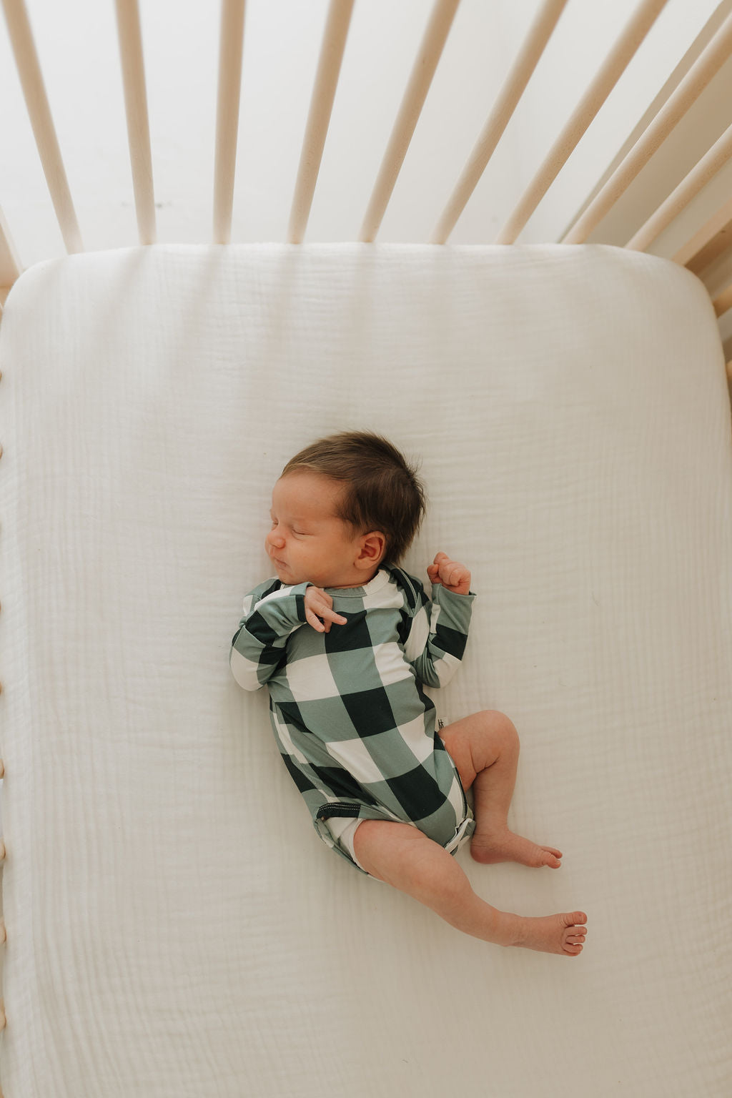 A sleeping newborn in a Long Sleeve Bamboo Snapsuit featuring the Evergreen Gingham pattern from forever french baby lies on a white crib mattress. The baby's arms are slightly bent, and they are positioned on their side with their legs relaxed. Soft light filters through the crib bars above.