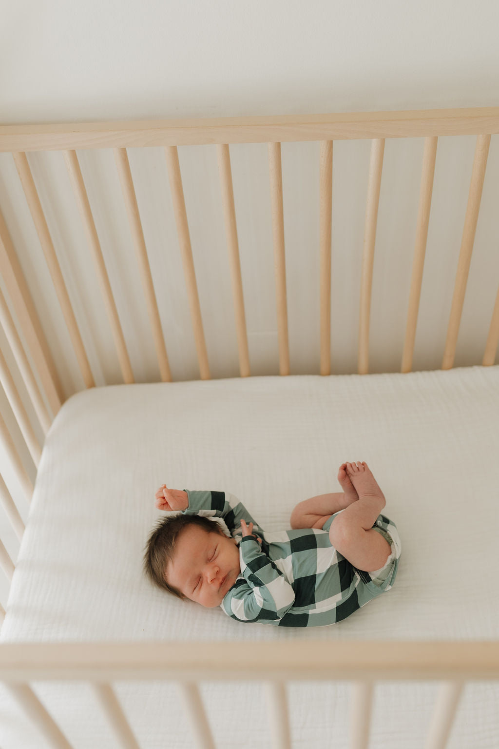 A baby peacefully sleeps in the Long Sleeve Bamboo Snapsuit in Evergreen Gingham by forever french baby, resting on a white mattress inside a wooden crib. The hypo-allergenic material of the clothing contributes to the tranquil scene, highlighted by gentle, natural lighting.