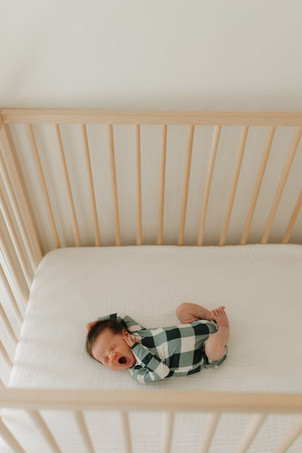 An infant dressed in the Evergreen Gingham Long Sleeve Bamboo Snapsuit from forever french baby yawns while lying on their back in a wooden crib with a white mattress. Crafted from bamboo fabric, this hypo-allergenic snapsuit ensures comfort for the little one.