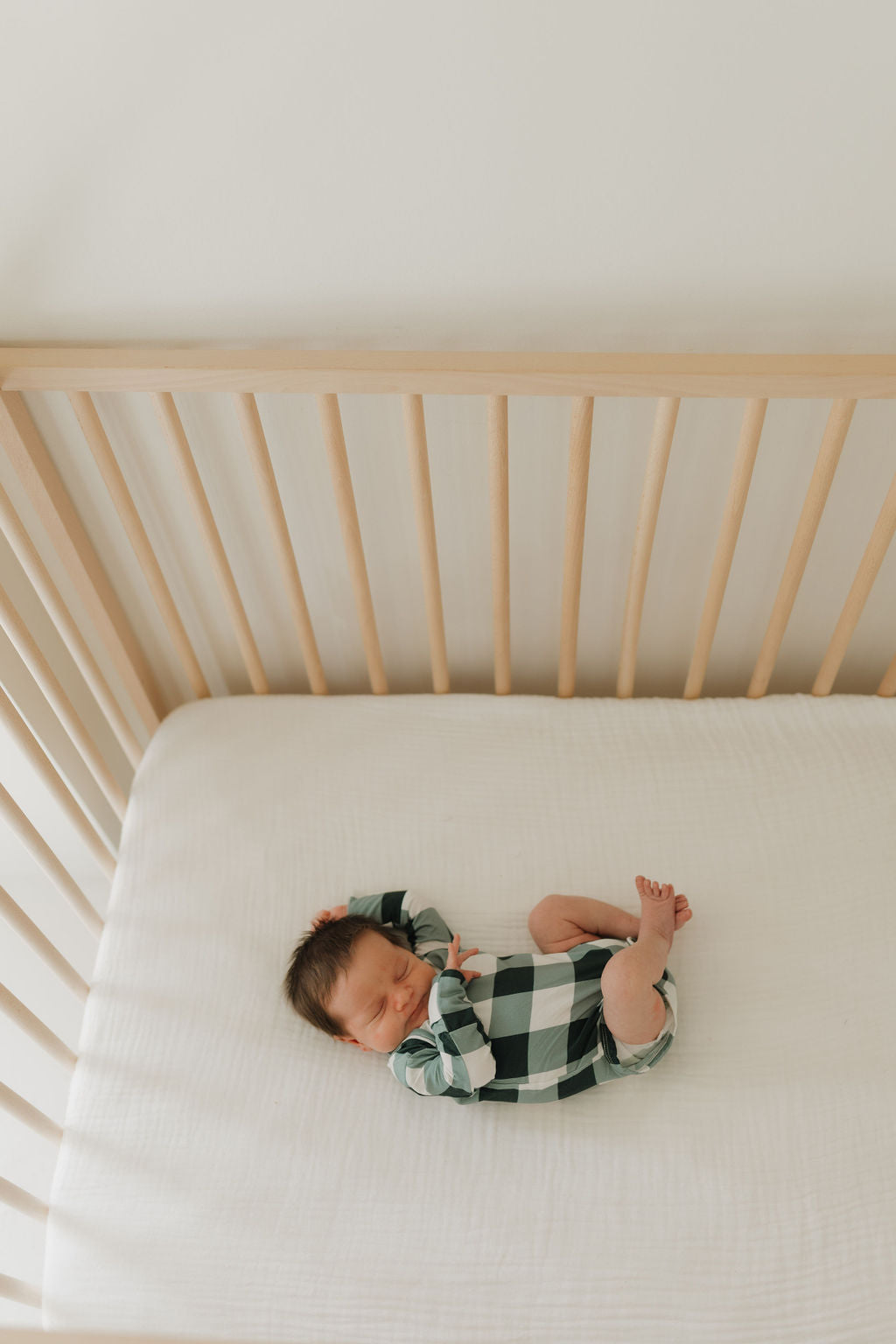 A baby wearing the Long Sleeve Bamboo Snapsuit in Evergreen Gingham by forever french baby sleeps soundly in a wooden crib on a breathable, hypo-allergenic white bamboo fabric mattress, set against a plain white background.