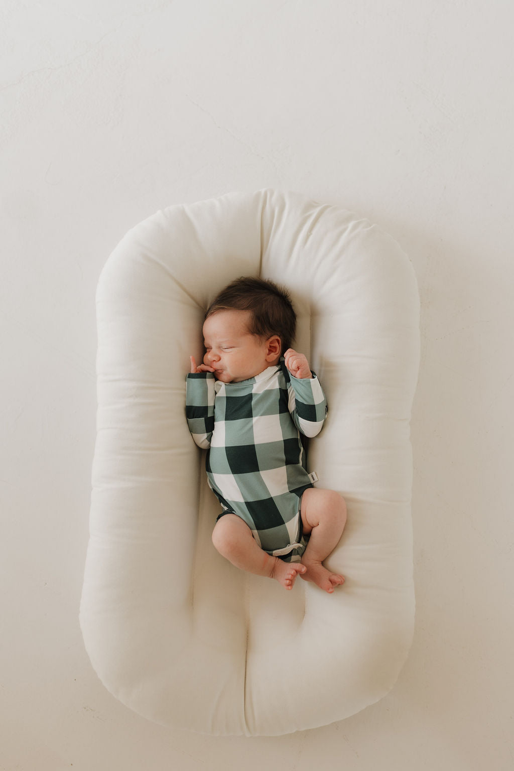 A baby is resting on a white, cushioned surface, wrapped in the Evergreen Gingham Long Sleeve Bamboo Snapsuit by forever french baby.