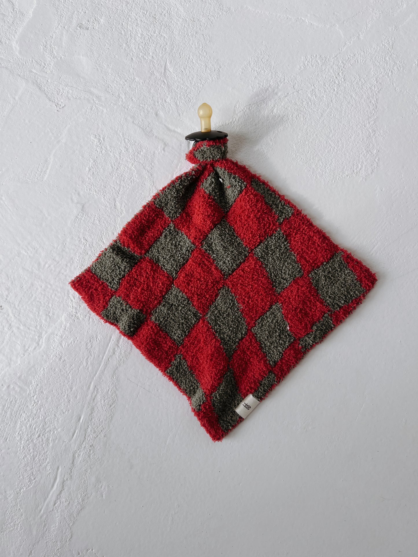 The "Lovey | Retro Christmas" blanket by forever french baby, crafted from soft microfiber feather yarn, features a checkered red and gray design with alternating diamond patterns. It hangs against a textured white wall with a wooden knob at the top.