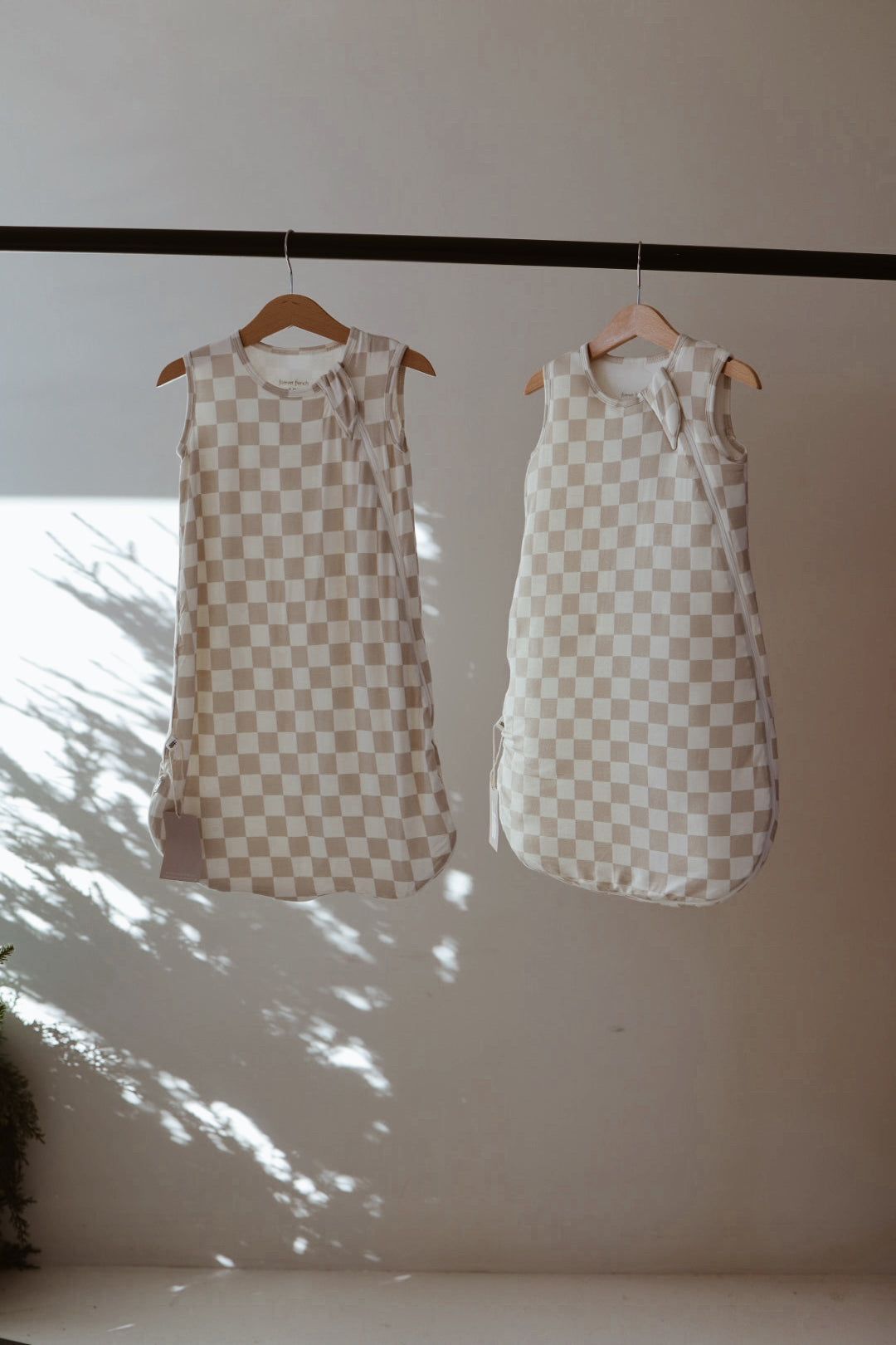 Two forever french baby Bamboo Sleep Sack | Checkerboard in beige and white hang on wooden hangers from a rod, with sunlight casting plant shadows on the wall behind them.