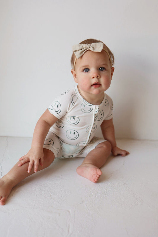 A baby with light hair wears the forever french baby Shortie Bamboo Zip in Green Smiles and a cream bow headband, sitting on a white floor against a white wall, looking upward in cozy, hypoallergenic clothing.