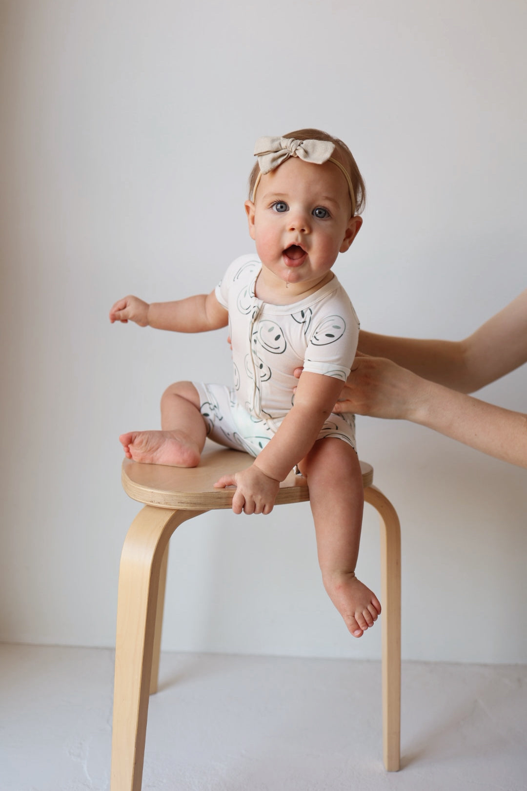 A baby in the forever french baby Shortie Bamboo Zip | Green Smiles onesie with a smiling face pattern and matching bow headband sits on a wooden stool, supported by an adult hand. The background is plain and light-colored.