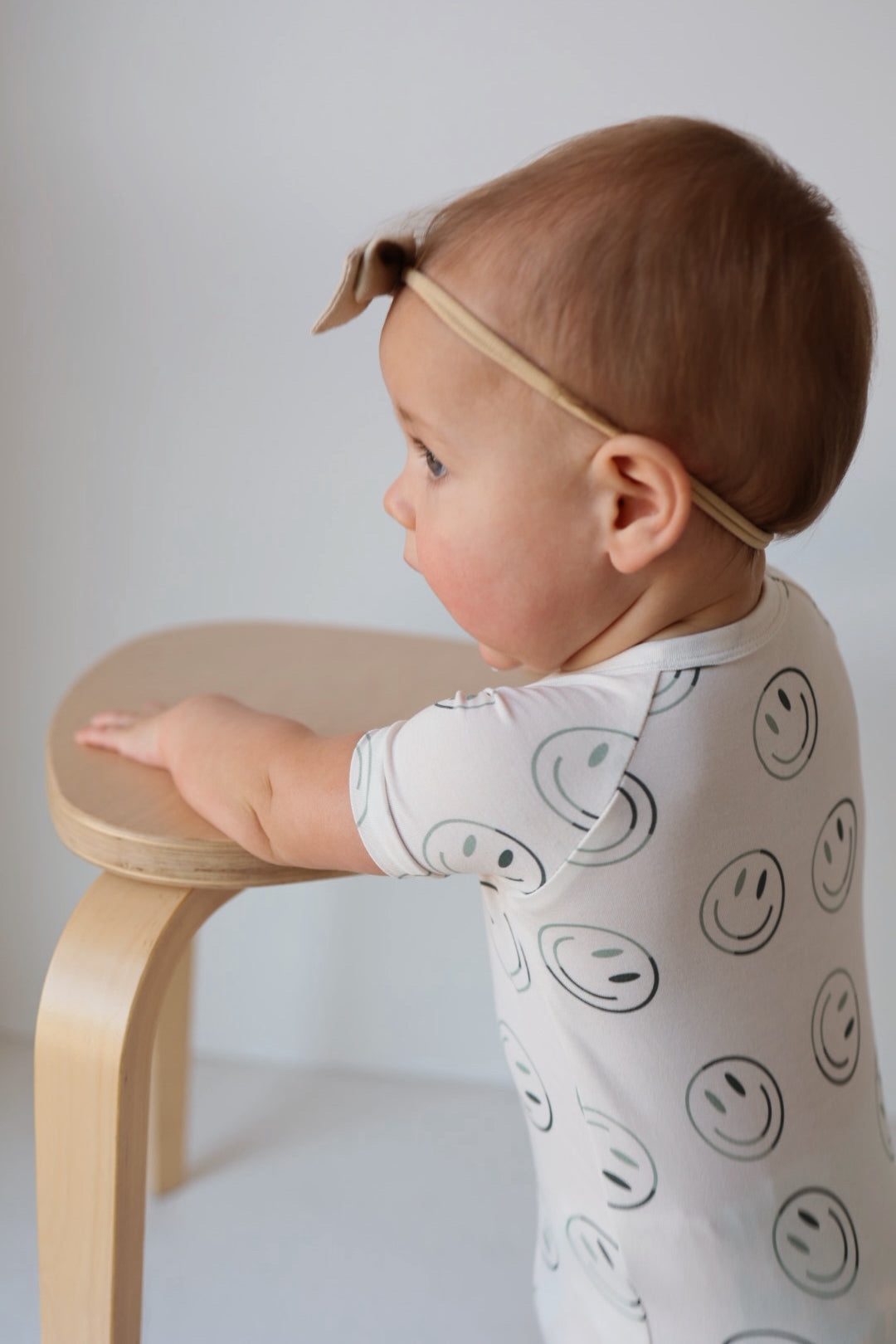 A baby in the forever french baby Shortie Bamboo Zip | Green Smiles stands sideways, holding a light wooden stool. The outfit features a white onesie with green smiley faces and a beige headband, set against a neutral background.