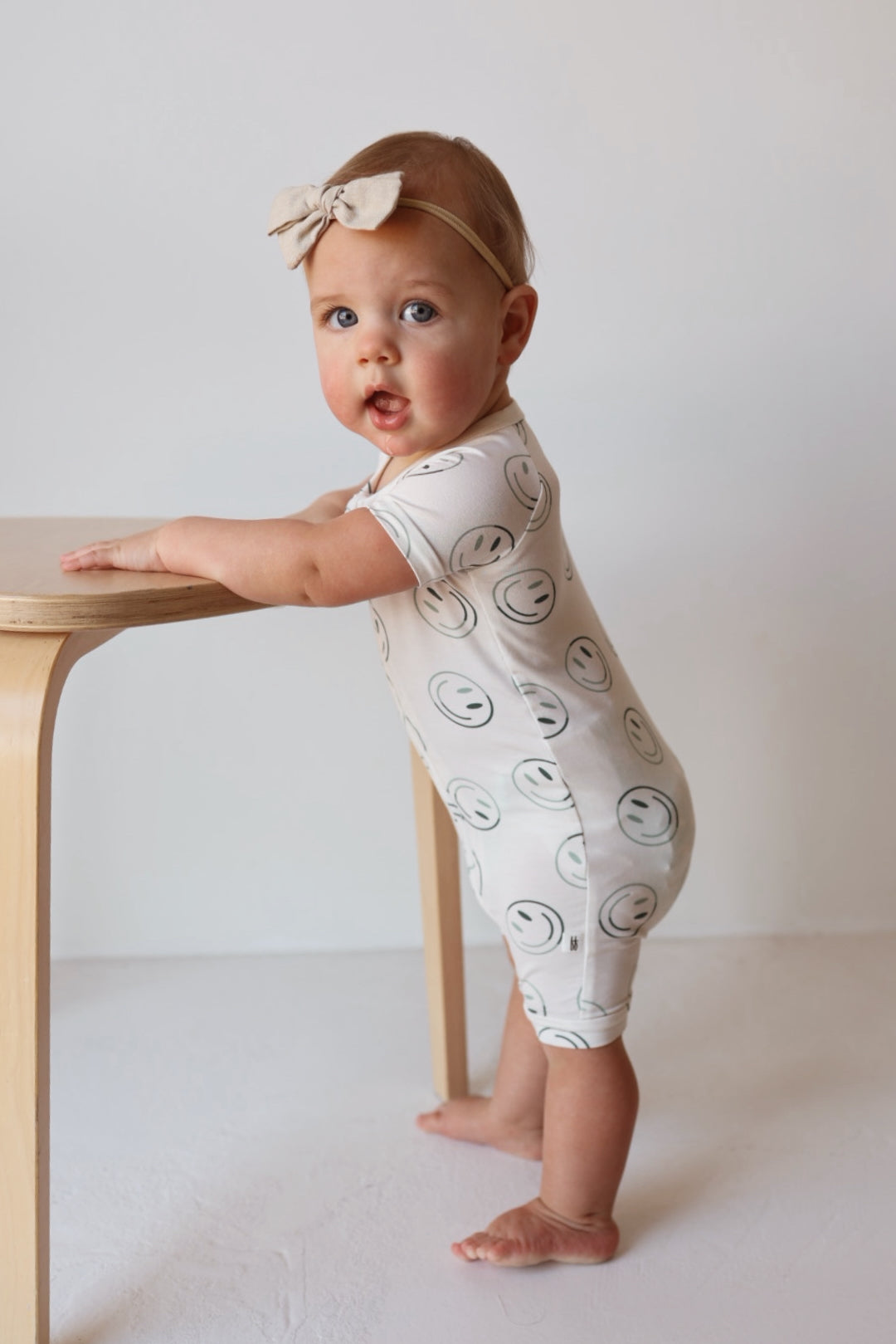 A baby in a forever french baby Shortie Bamboo Zip | Green Smiles onesie with smiley faces and a matching headband stands holding a light wooden table, looking at the camera.