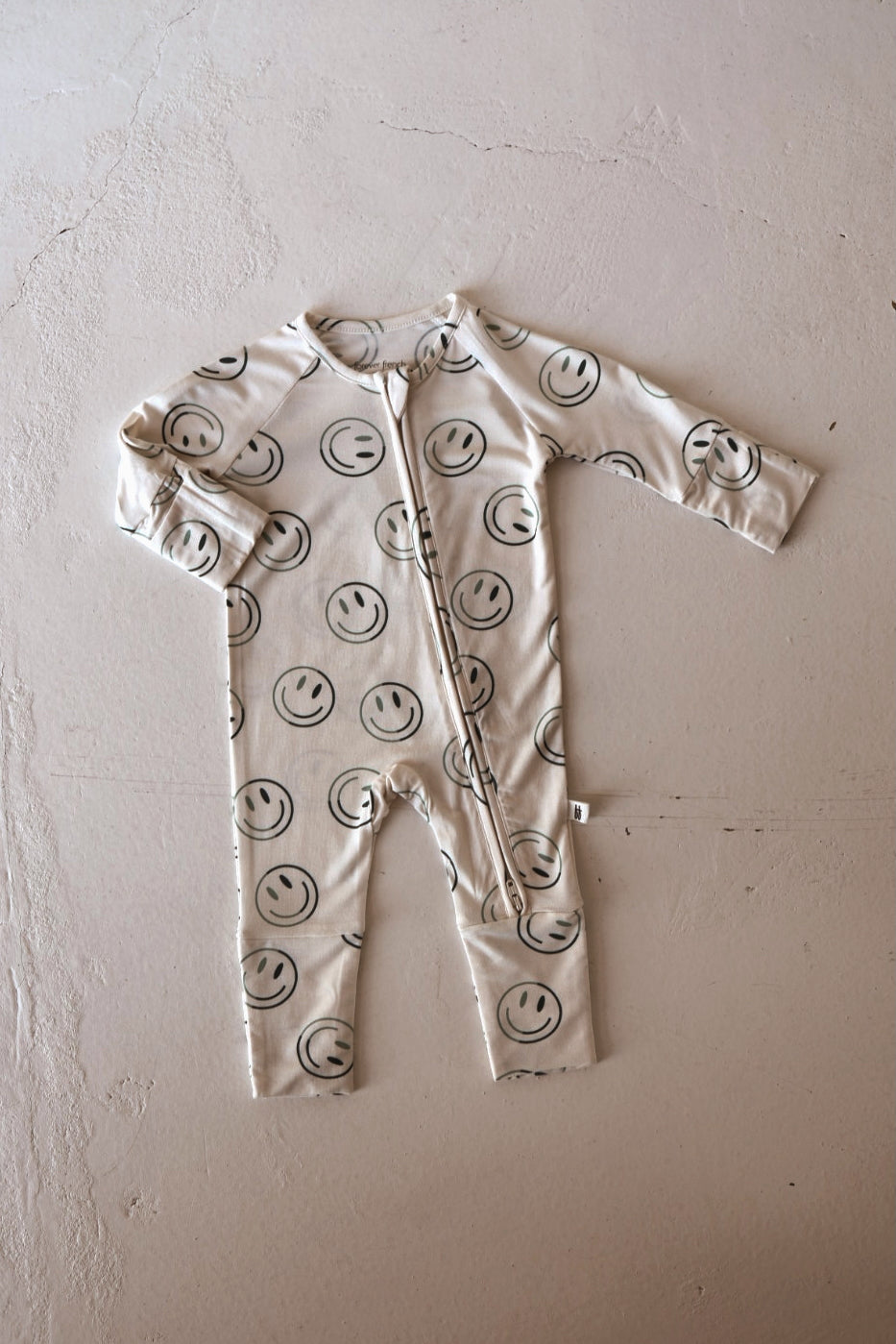 The forever french baby Bamboo Zip Pajama | Green Smiles is a cream onesie with black smiley faces, made from breathable bamboo, featuring long sleeves and legs plus a front zipper. The onesie appears laid flat on a light textured surface.