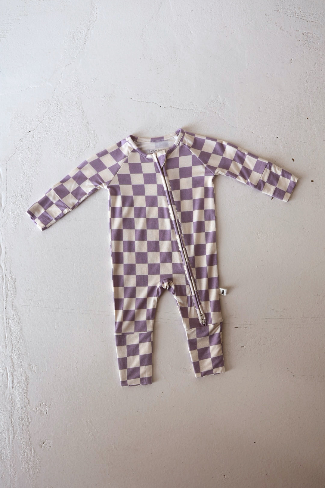 The forever french baby Bamboo Zip Pajama in Grape Soda, a long-sleeve breathable romper with a lavender and cream checkerboard pattern and diagonal front zipper, is displayed flat on a light-textured surface.