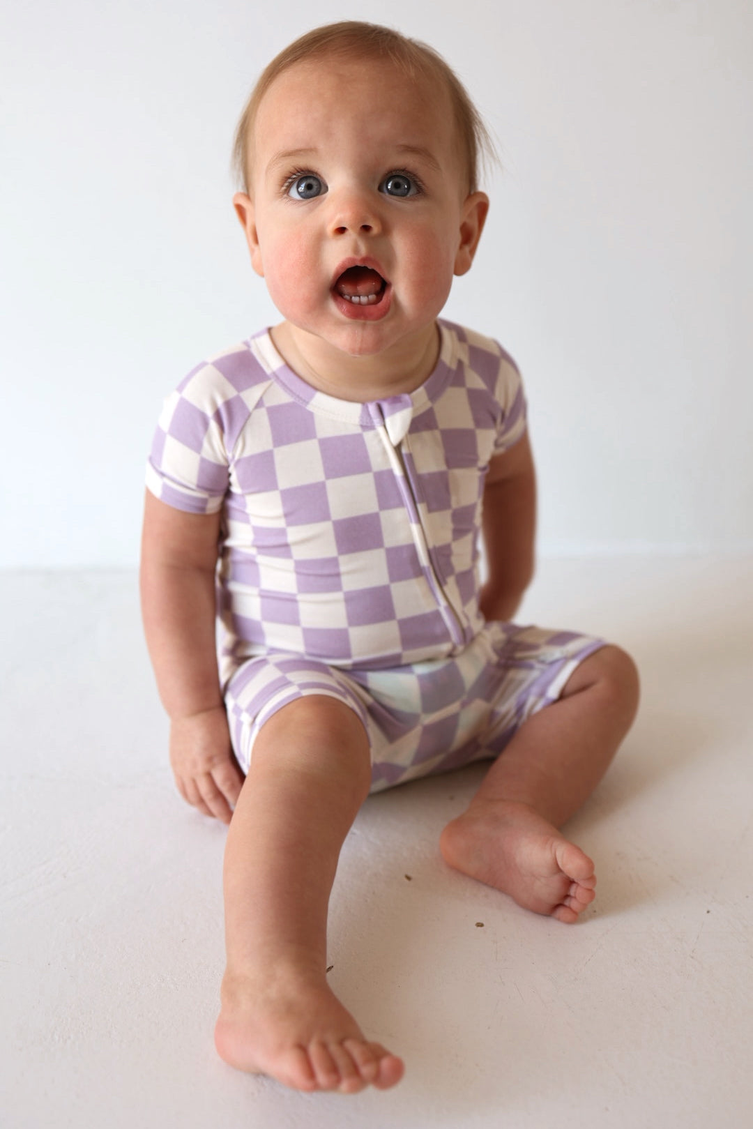 A baby in the forever french baby Shortie Bamboo Zip | Grape Soda romper sits on a white floor, one leg out, mouth open, and gazing up with wide blue eyes.