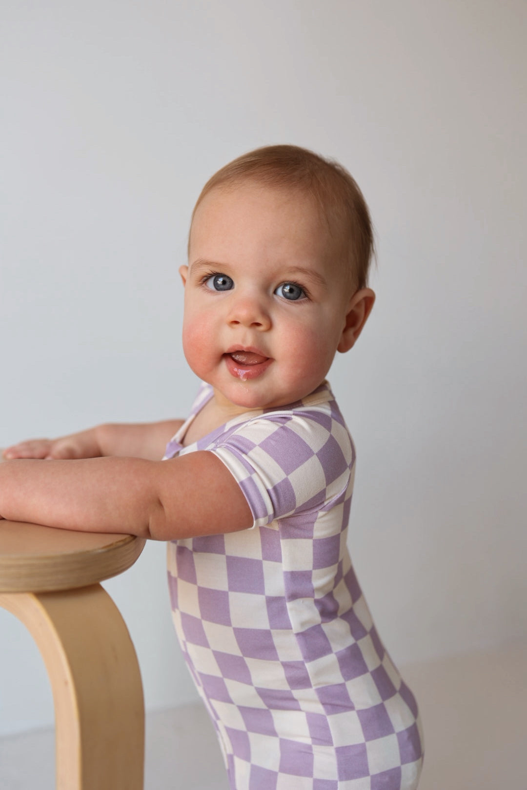 A smiling baby with light brown hair and blue eyes stands holding a wooden stool, wearing the forever french baby Shortie Bamboo Zip in Grape Soda—a hypoallergenic romper that’s gentle on skin—posed against a plain backdrop.