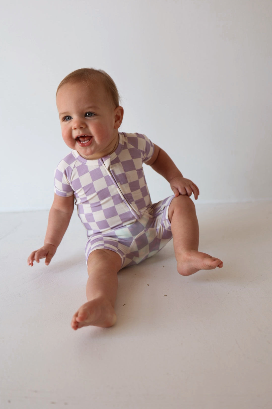 A smiling baby sits on the floor wearing the forever french baby Shortie Bamboo Zip in Grape Soda, a short-sleeve purple and white checkered romper, against a plain white background.