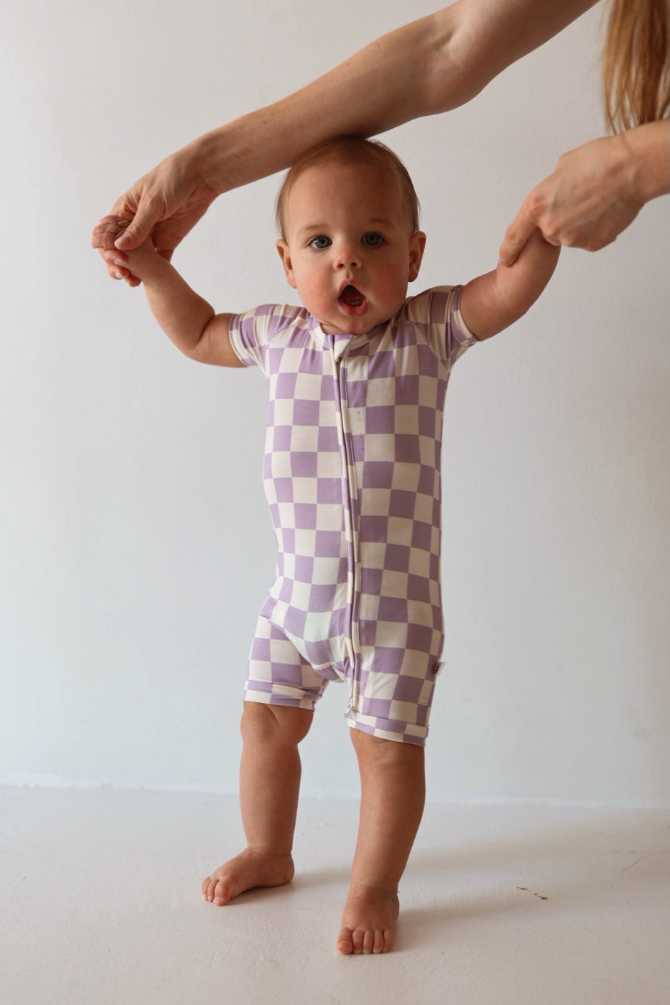 A baby in the forever french baby Shortie Bamboo Zip | Grape Soda romper stands supported by an adult, looking ahead with an open-mouthed expression against a plain, light background.