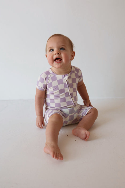 A baby in the forever french baby Shortie Bamboo Zip | Grape Soda romper sits barefoot on a light-colored floor, gazing up with an open-mouthed smile against a plain background.