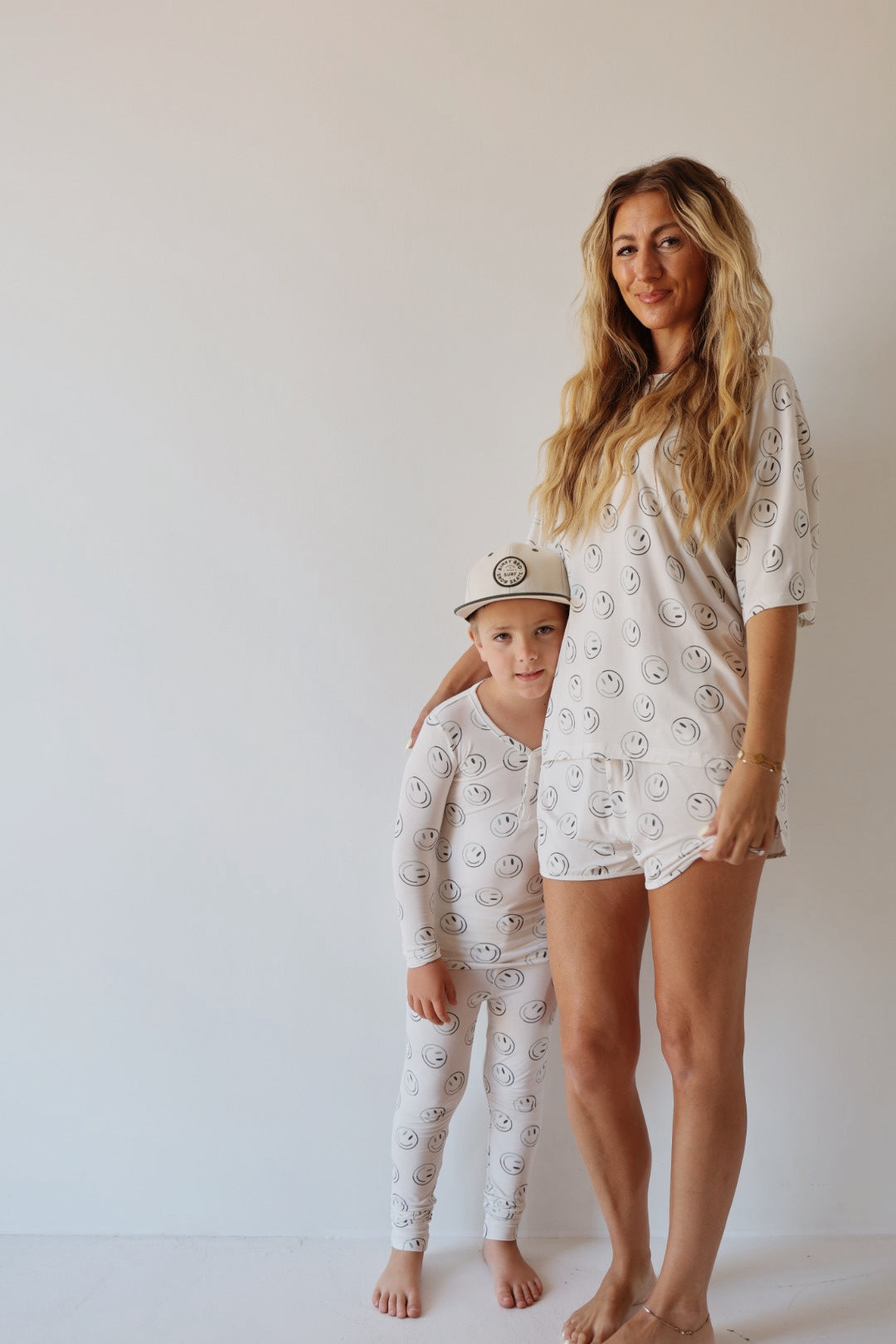 A woman and child smile side by side in forever french baby Bamboo Two Piece Pajamas—Green Smiles, featuring a cheerful pattern; the child also wears a white hat with a circular logo against a plain, light background.