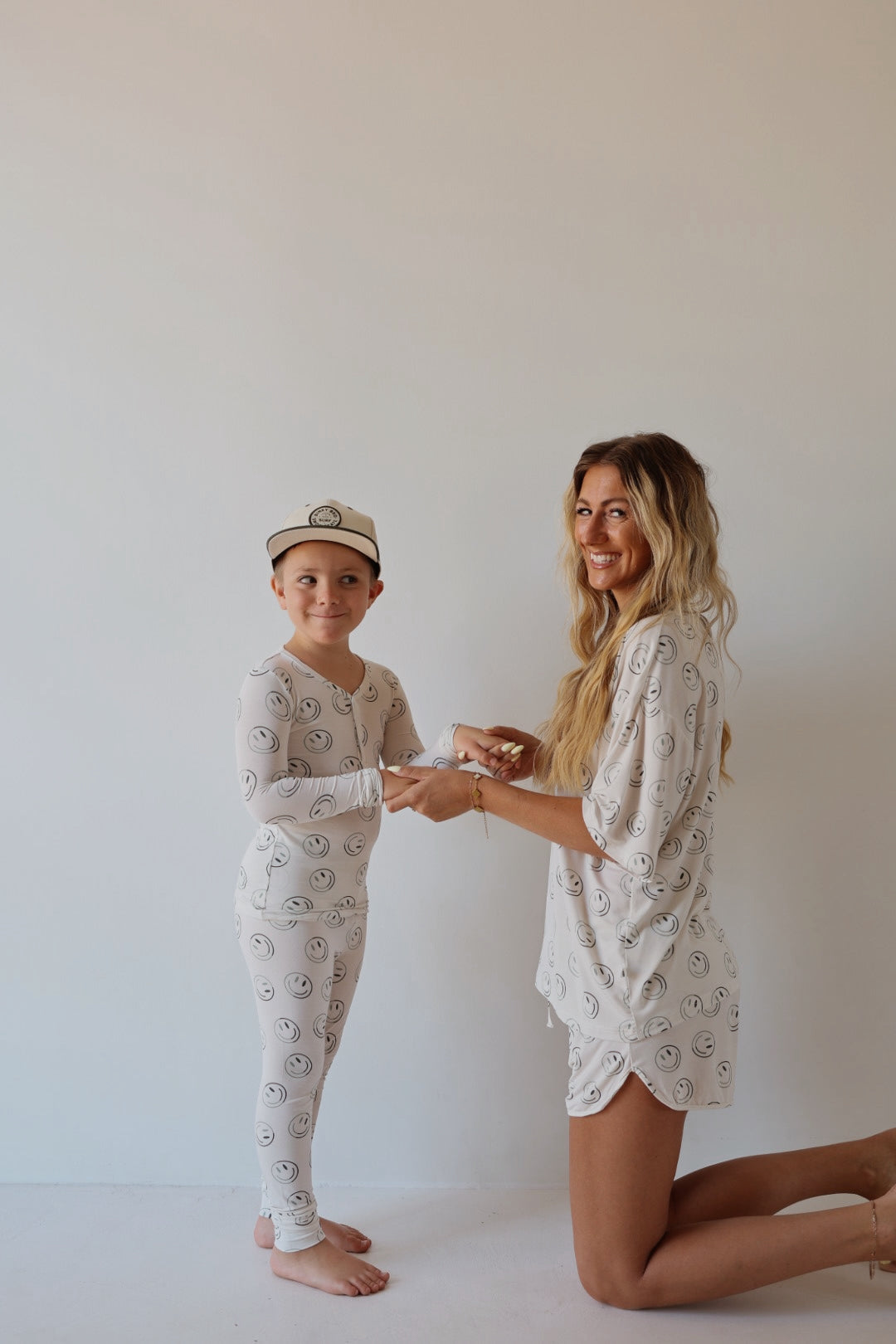 A woman kneels beside a smiling child, both dressed in forever french baby’s Bamboo Two Piece Pajamas | Green Smiles, featuring a cheerful pattern. The child wears a white cap as they hold hands against a plain light background.