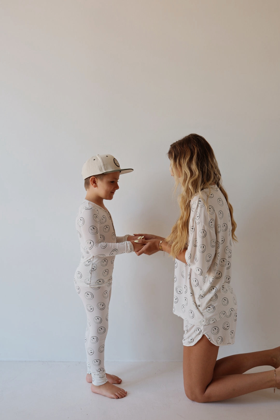 A woman kneels and holds hands with a smiling boy, both wearing forever french baby’s Bamboo Two Piece Pajamas | Green Smiles—matching white bamboo sleepwear with smiley faces, shown against a plain white wall.