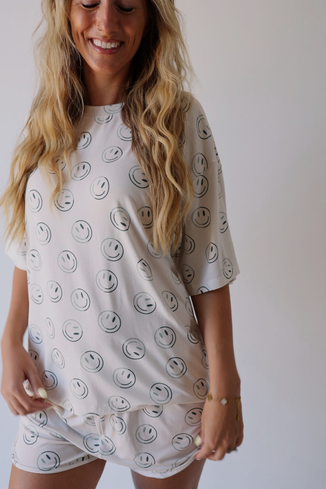 Woman with long blonde hair wears the forever french baby Women's Bamboo Short Set in Green Smiles, featuring black smiley face prints. She stands against a plain background, smiling with her head slightly tilted down.