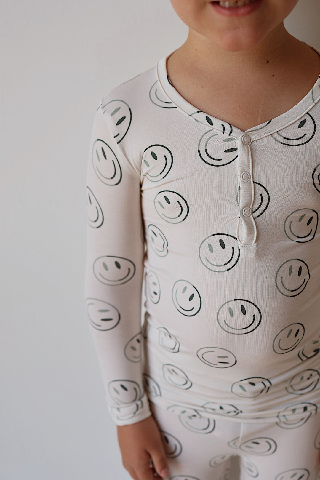 A child in forever french baby Bamboo Two Piece Pajamas—Green Smiles, featuring a cheerful black smiley face print, stands against a white background with only the lower half of their face and torso visible.